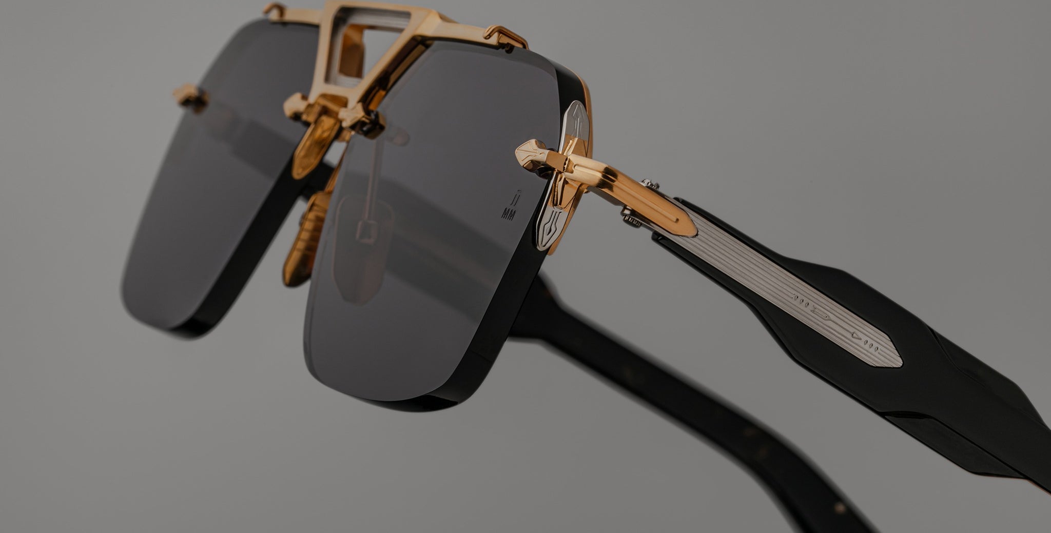 Close-up of Jacques Marie Mage Silverton sunglasses with dark rectangular lenses, gold metal frame, and black arms with metallic detailing, set against a plain gray background.