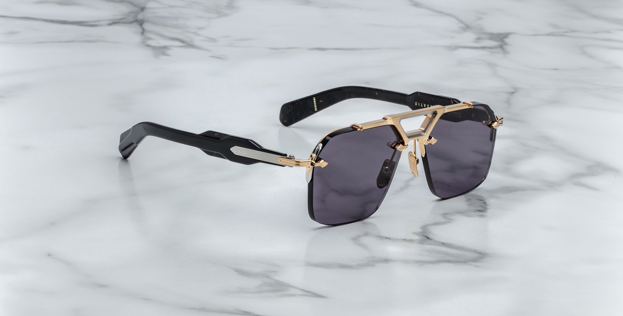 The Jacques Marie Mage Silverton sunglasses feature dark rectangular lenses, gold-tone metal frames, and thick black arms, displayed on a white marble surface with grey veining.