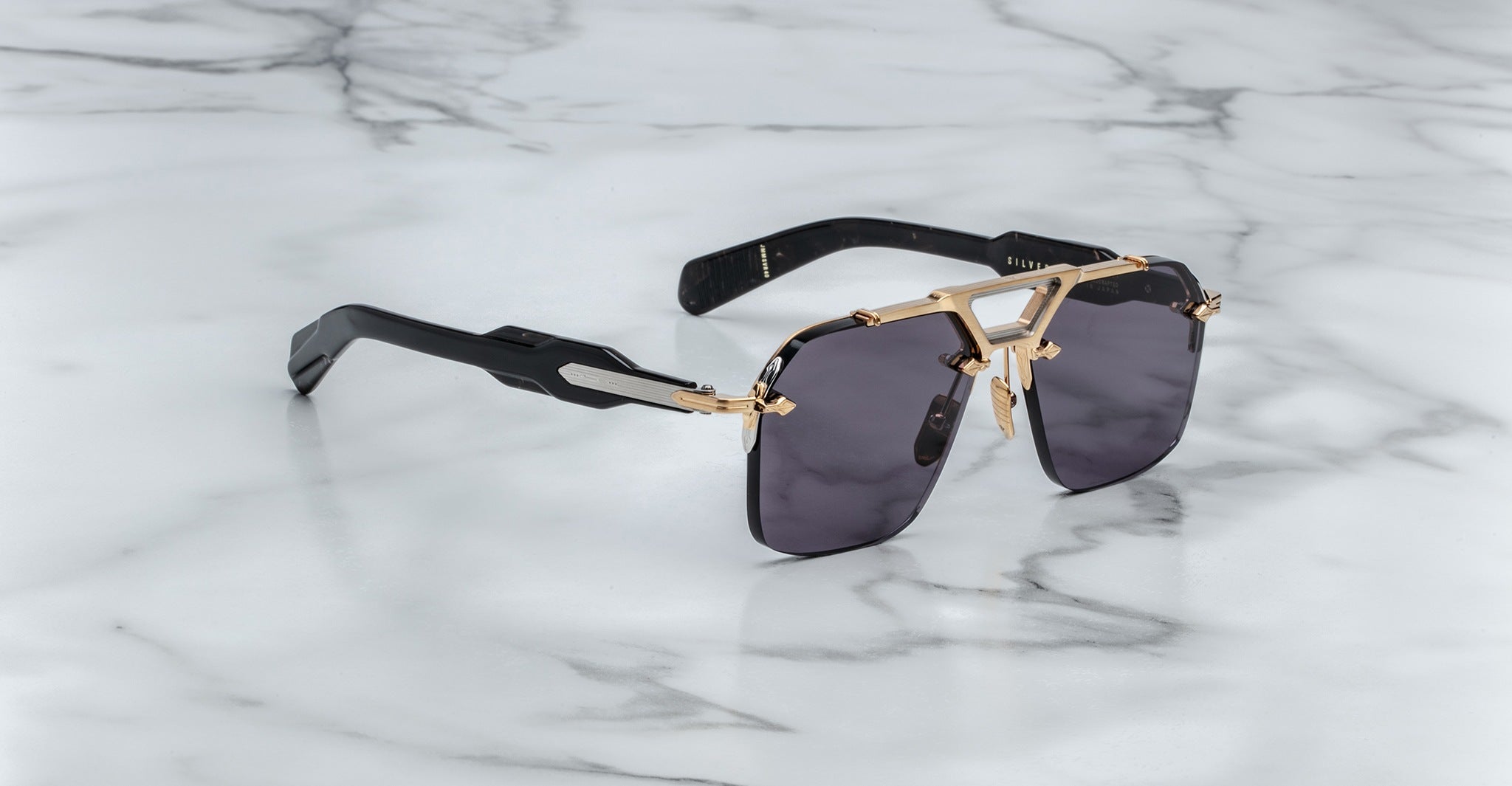 The Jacques Marie Mage Silverton sunglasses feature dark rectangular lenses, gold-tone metal frames, and thick black arms, displayed on a white marble surface with grey veining.