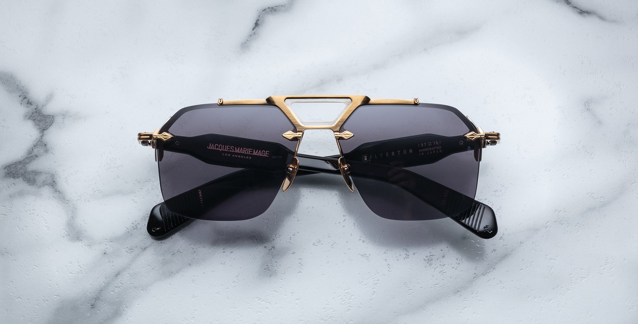 The Jacques Marie Mage Silverton sunglasses, featuring dark lenses and a gold metal geometric frame, are displayed on a white marble surface.