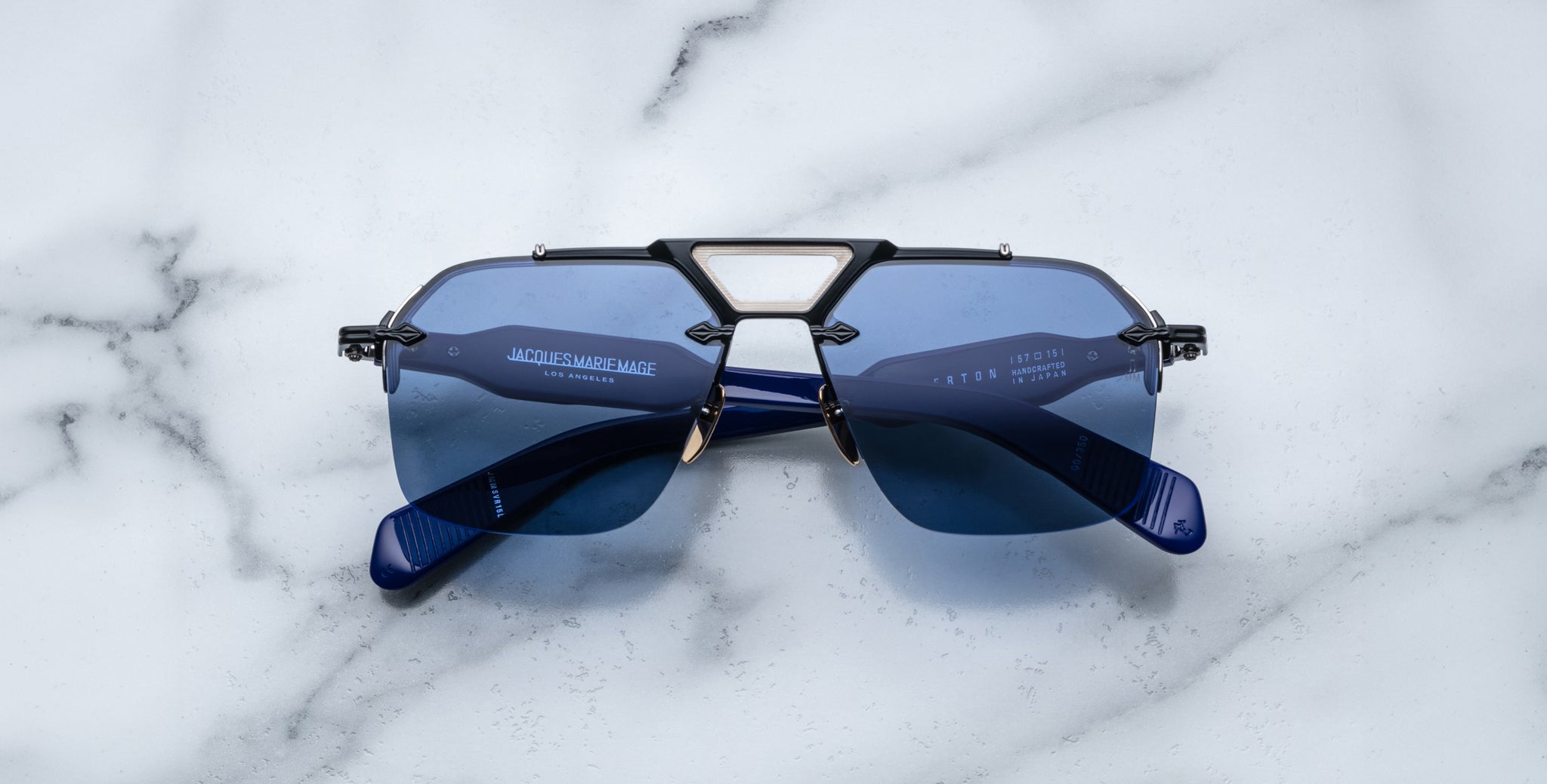 A pair of Jacques Marie Mage Silverton sunglasses with blue lenses and geometric metal accents on the bridge rests upside down on a white marble surface.