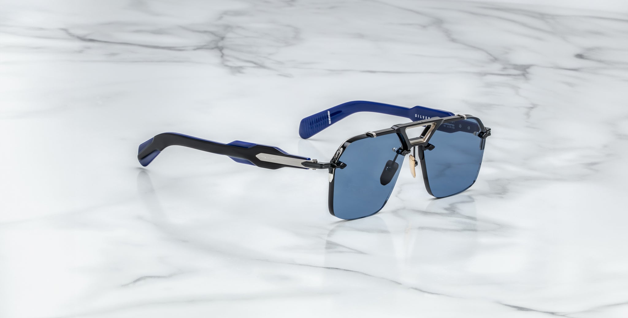 The Jacques Marie Mage Silverton sunglasses, featuring blue lenses, a metallic frame, and blue-black temples, are displayed on a white marble surface.