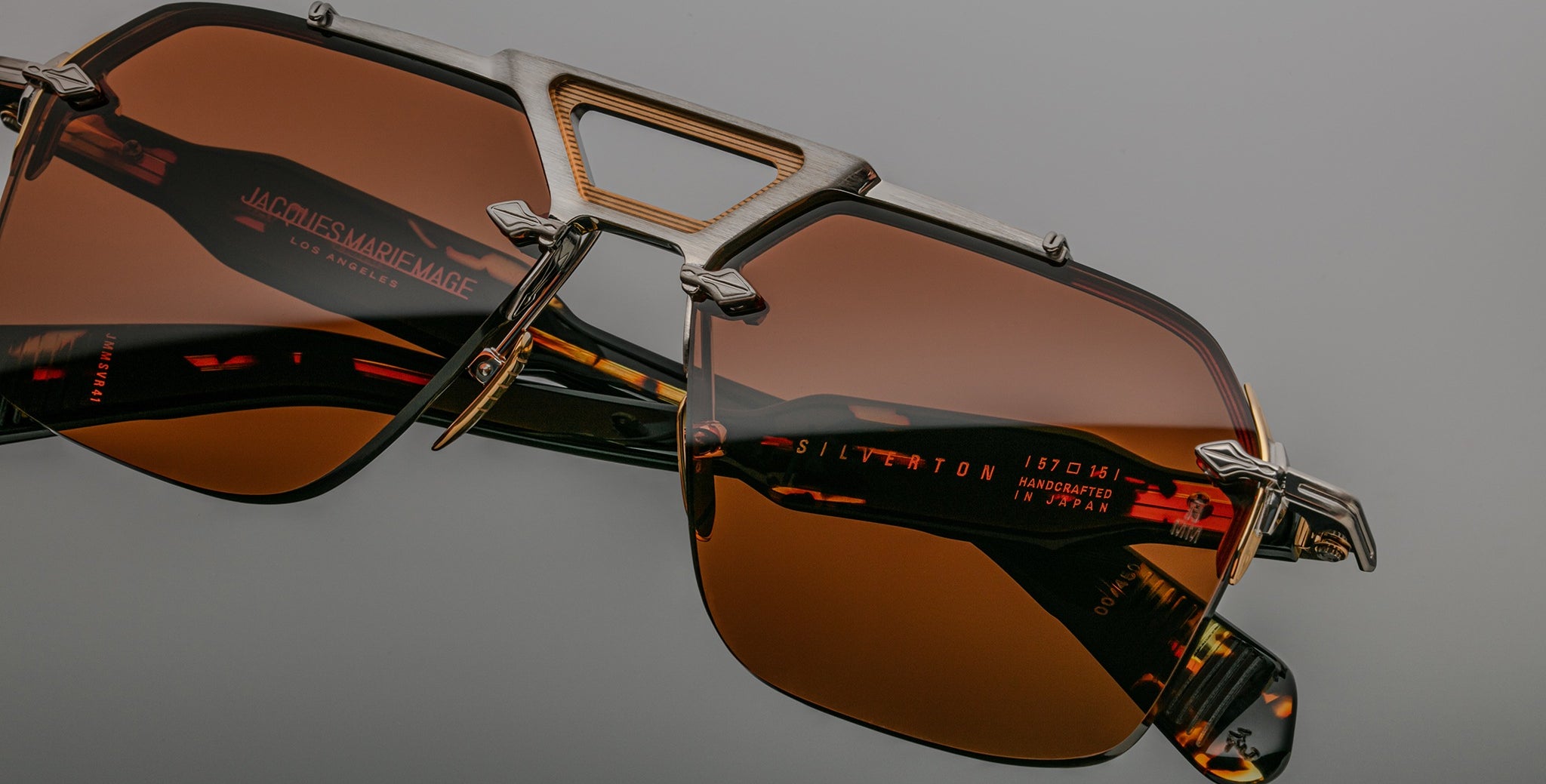 Close-up of Jacques Marie Mage Silverton sunglasses featuring brown tinted lenses, a unique geometric metal frame, and red text branding with specifications on the inner arm. Set against a neutral gray background.