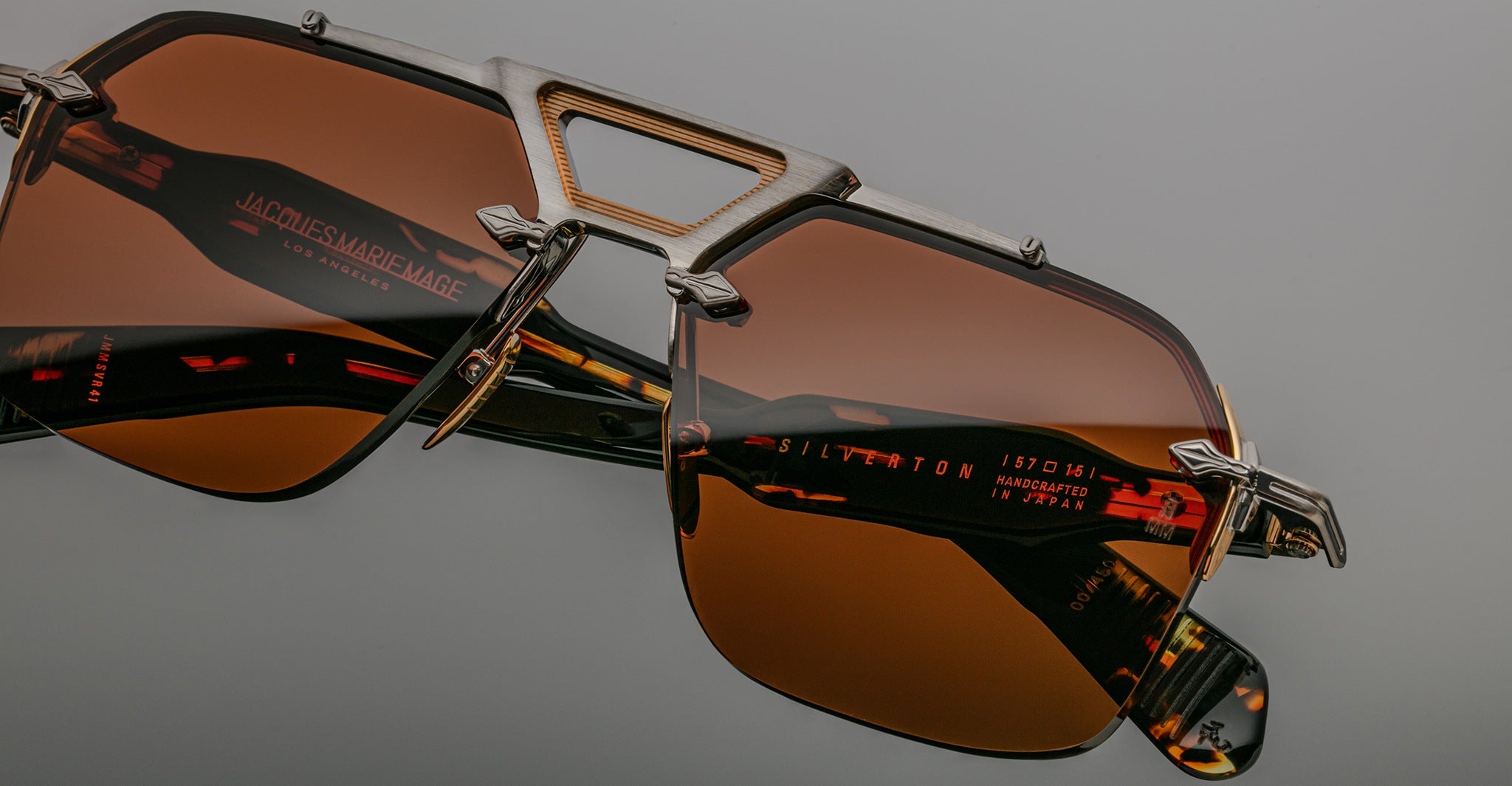 Close-up of Jacques Marie Mage Silverton sunglasses featuring brown tinted lenses, a unique geometric metal frame, and red text branding with specifications on the inner arm. Set against a neutral gray background.