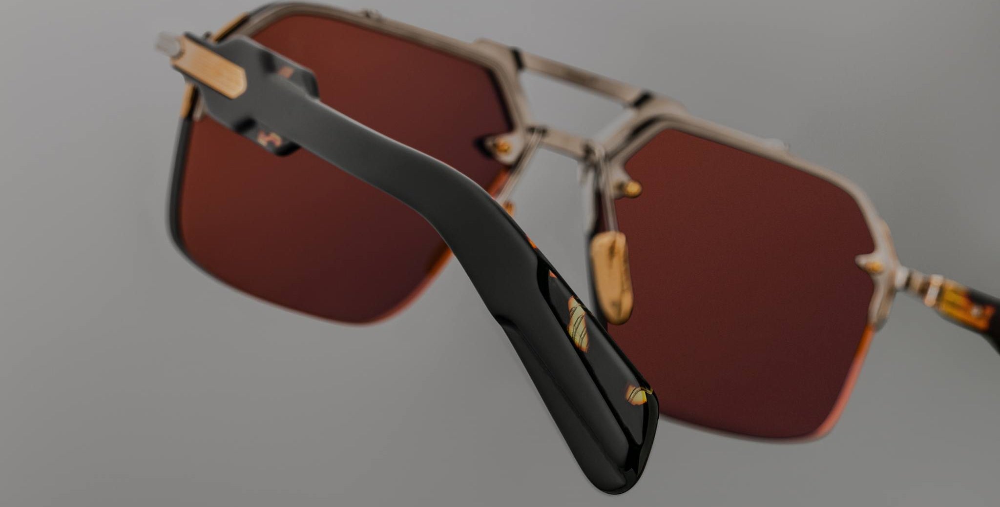 Close-up view of Jacques Marie Mage Silverton sunglasses featuring large hexagonal reddish-brown lenses with gold accents. A black temple arm is in focus, while the rest of the frame fades against a neutral background.