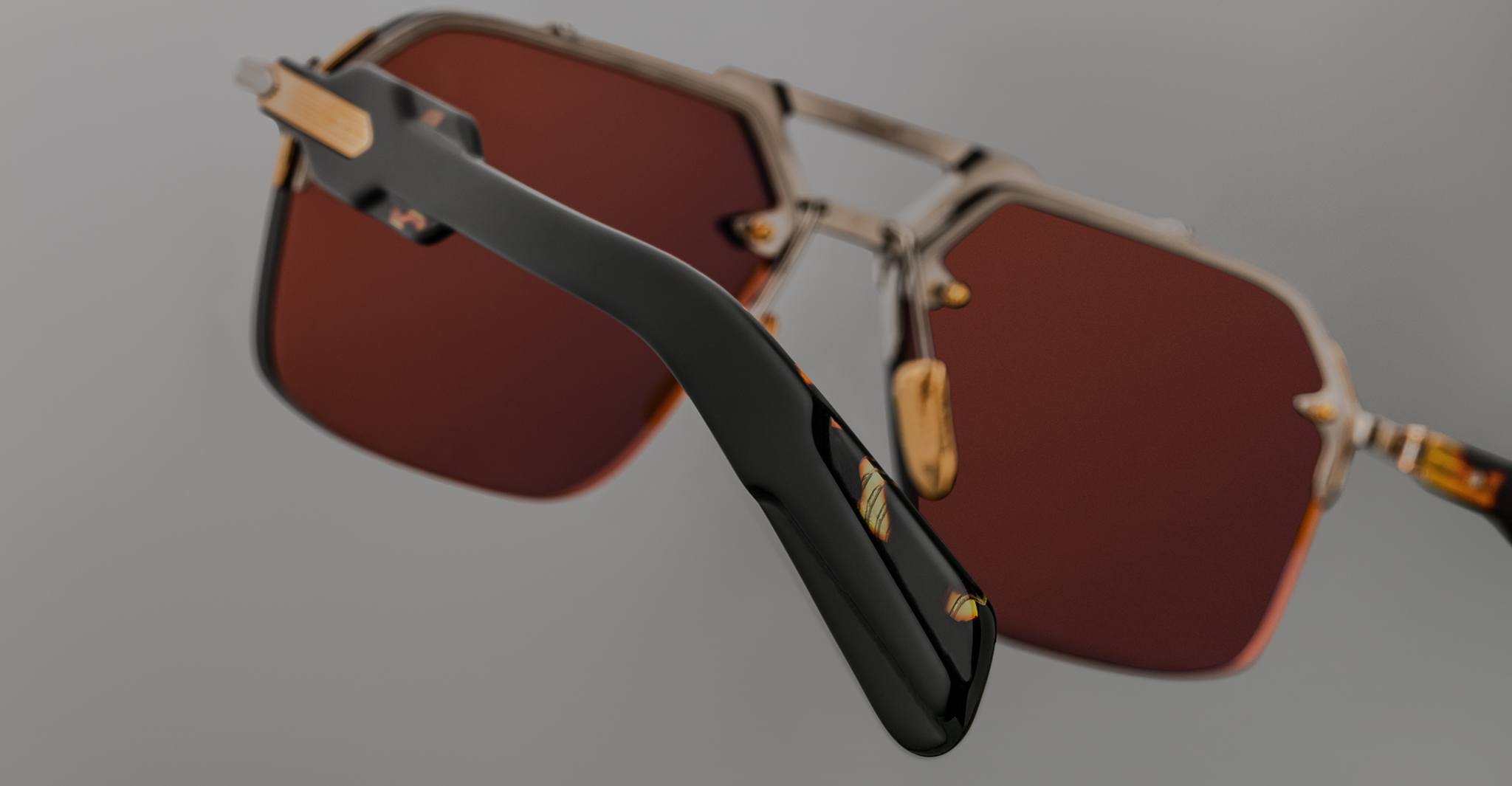 Close-up view of Jacques Marie Mage Silverton sunglasses featuring large hexagonal reddish-brown lenses with gold accents. A black temple arm is in focus, while the rest of the frame fades against a neutral background.