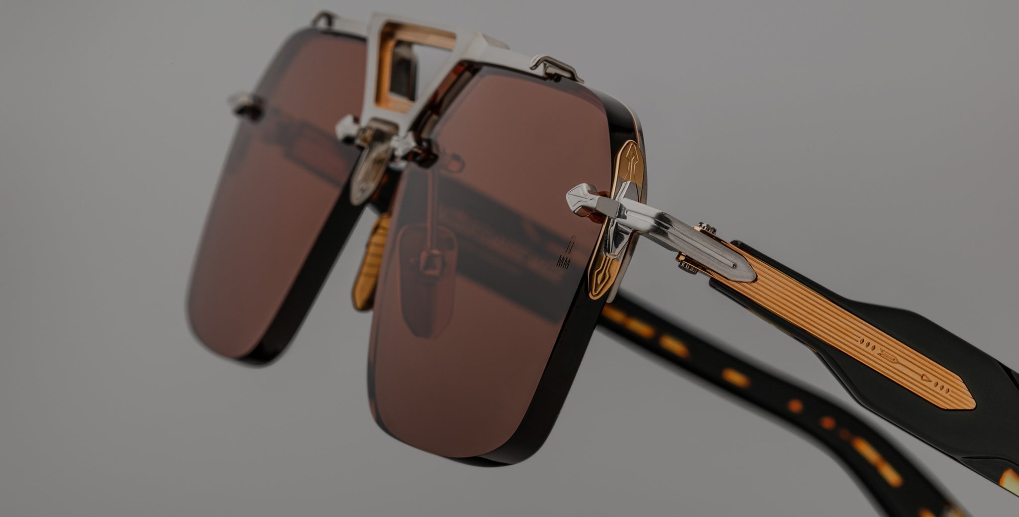 Close-up view of Jacques Marie Mage Silverton sunglasses featuring brown rectangular lenses, gold accents, and patterned black arms, set against a neutral background.