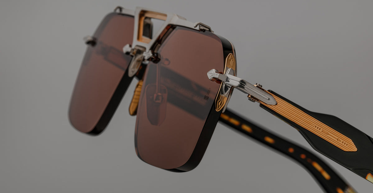 Close-up view of Jacques Marie Mage Silverton sunglasses featuring brown rectangular lenses, gold accents, and patterned black arms, set against a neutral background.
