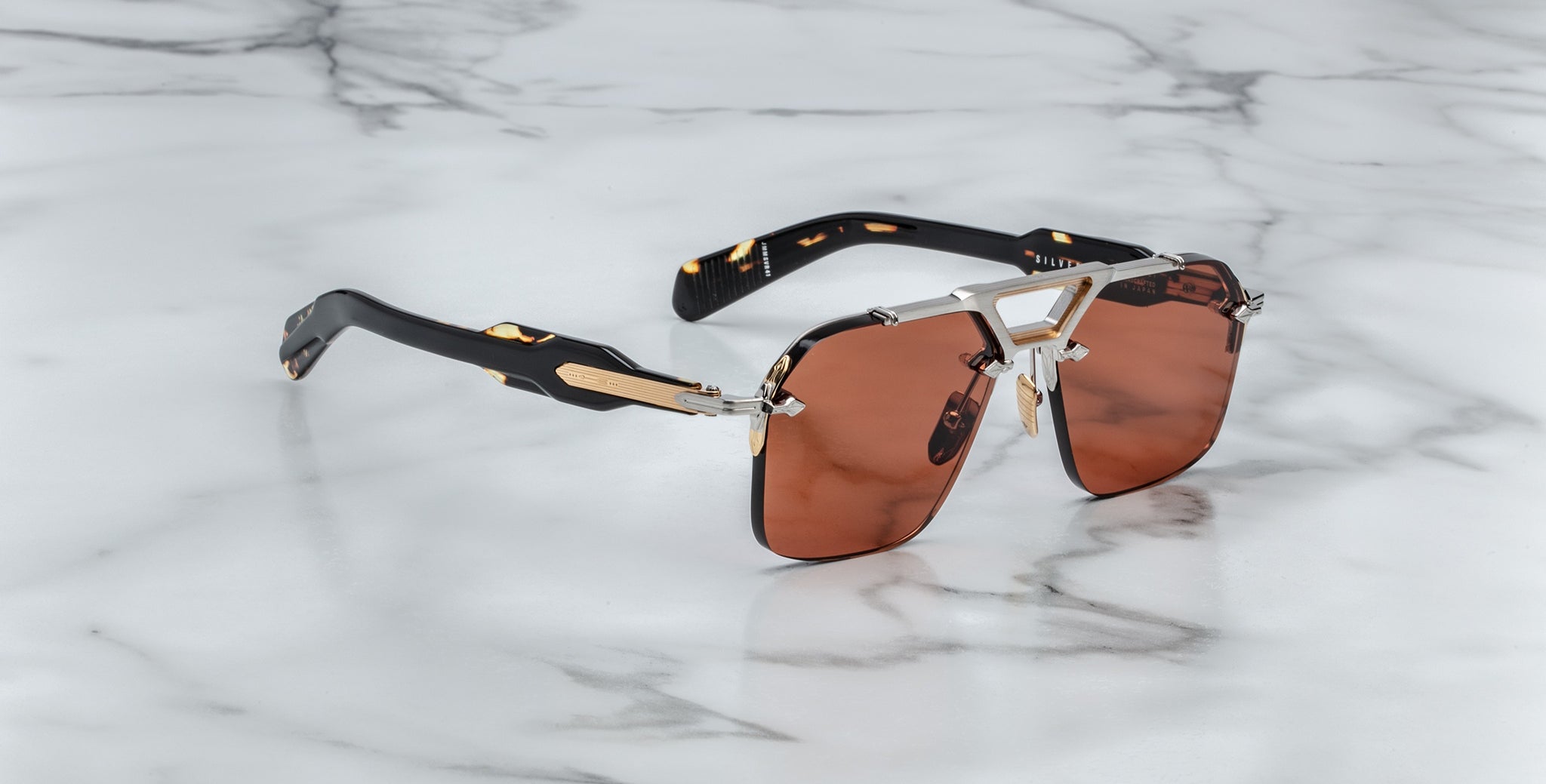 The Jacques Marie Mage Silverton sunglasses feature dark brown tinted lenses, gold and silver metal accents, and black arms, displayed on a white marble surface.
