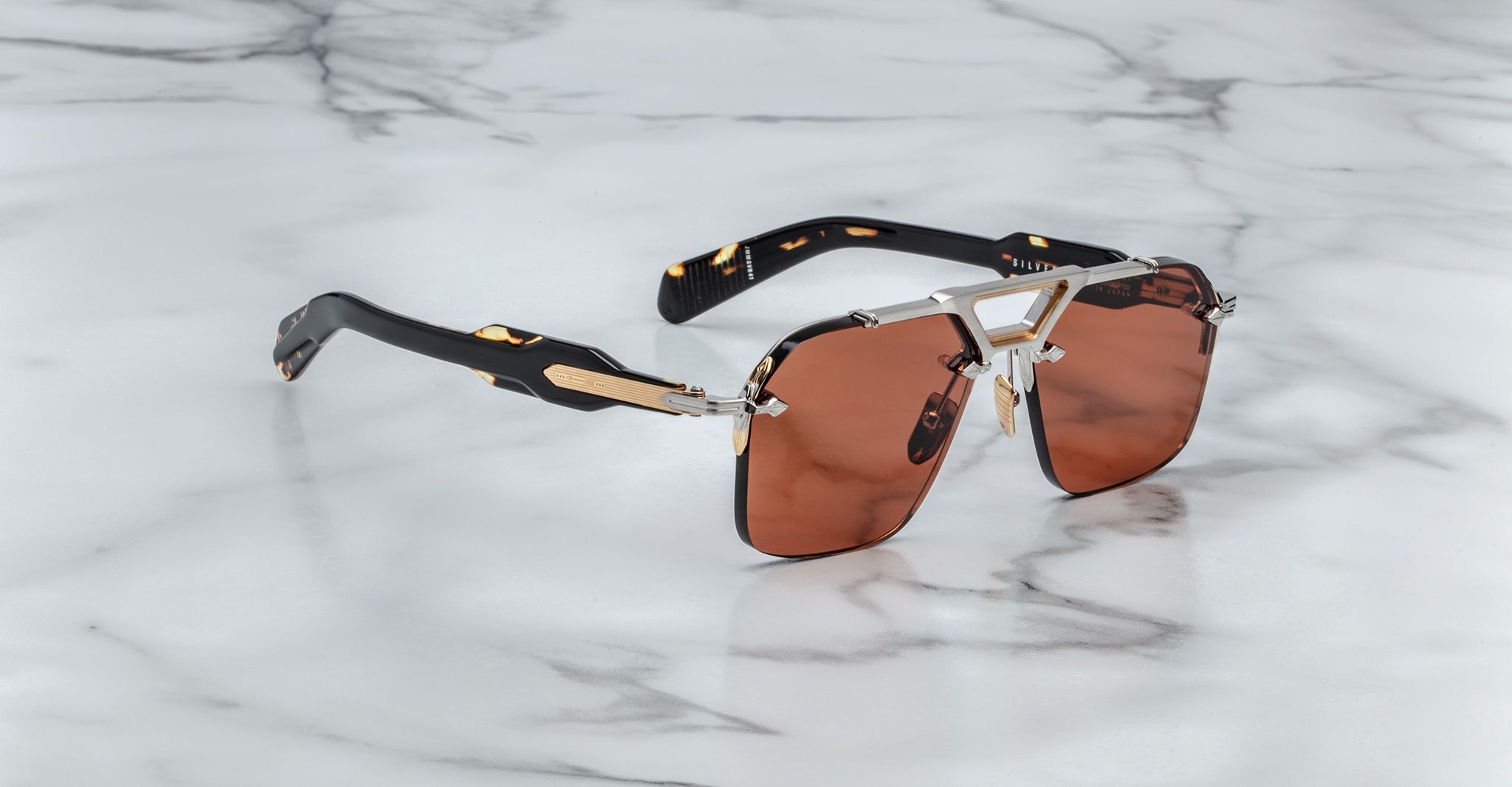 The Jacques Marie Mage Silverton sunglasses feature dark brown tinted lenses, gold and silver metal accents, and black arms, displayed on a white marble surface.