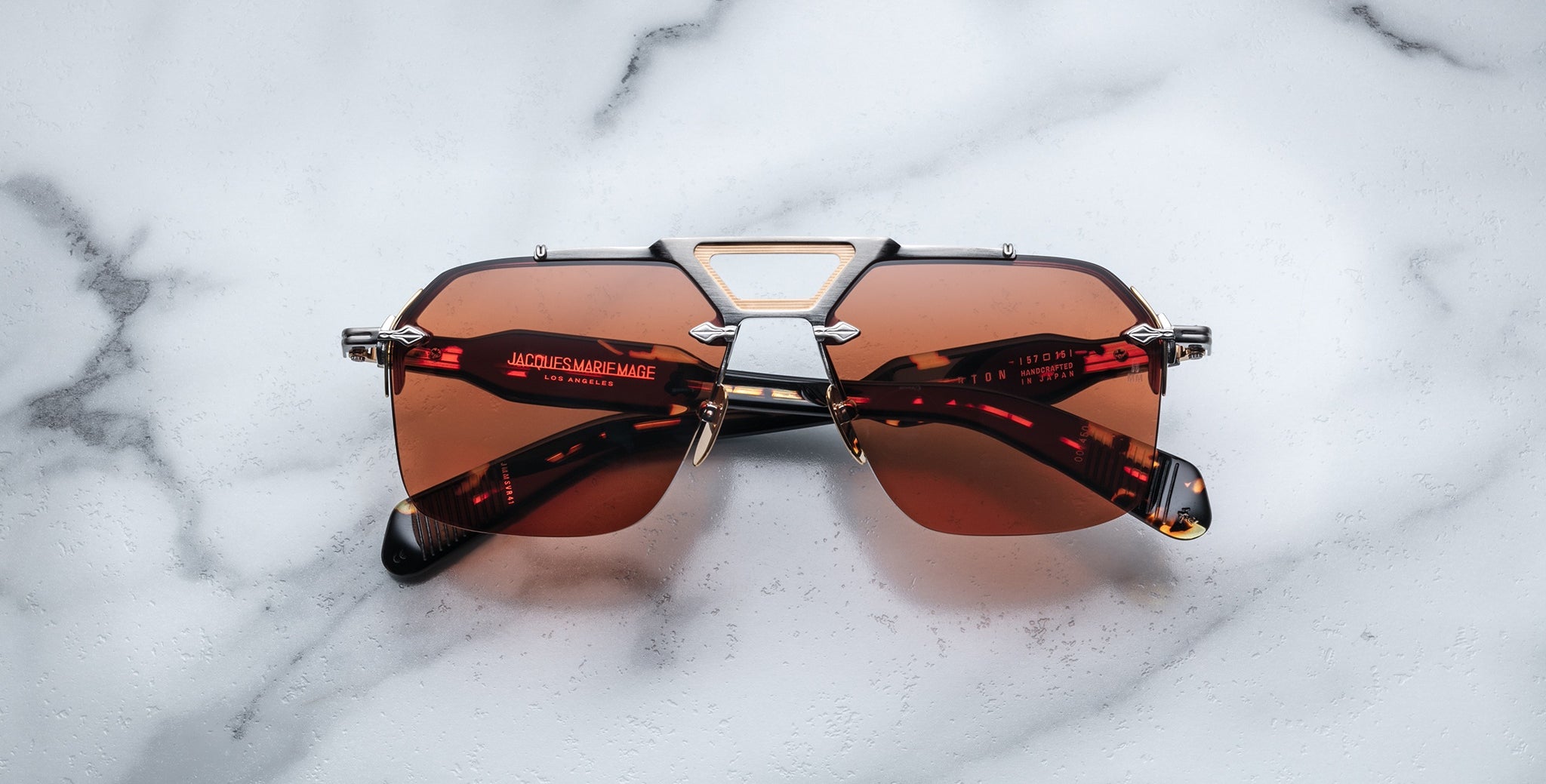 The Jacques Marie Mage Silverton rectangular sunglasses, featuring brown tinted lenses and a silver metal frame, rest on a white marble surface. The brand name and details appear in red text on the arms.