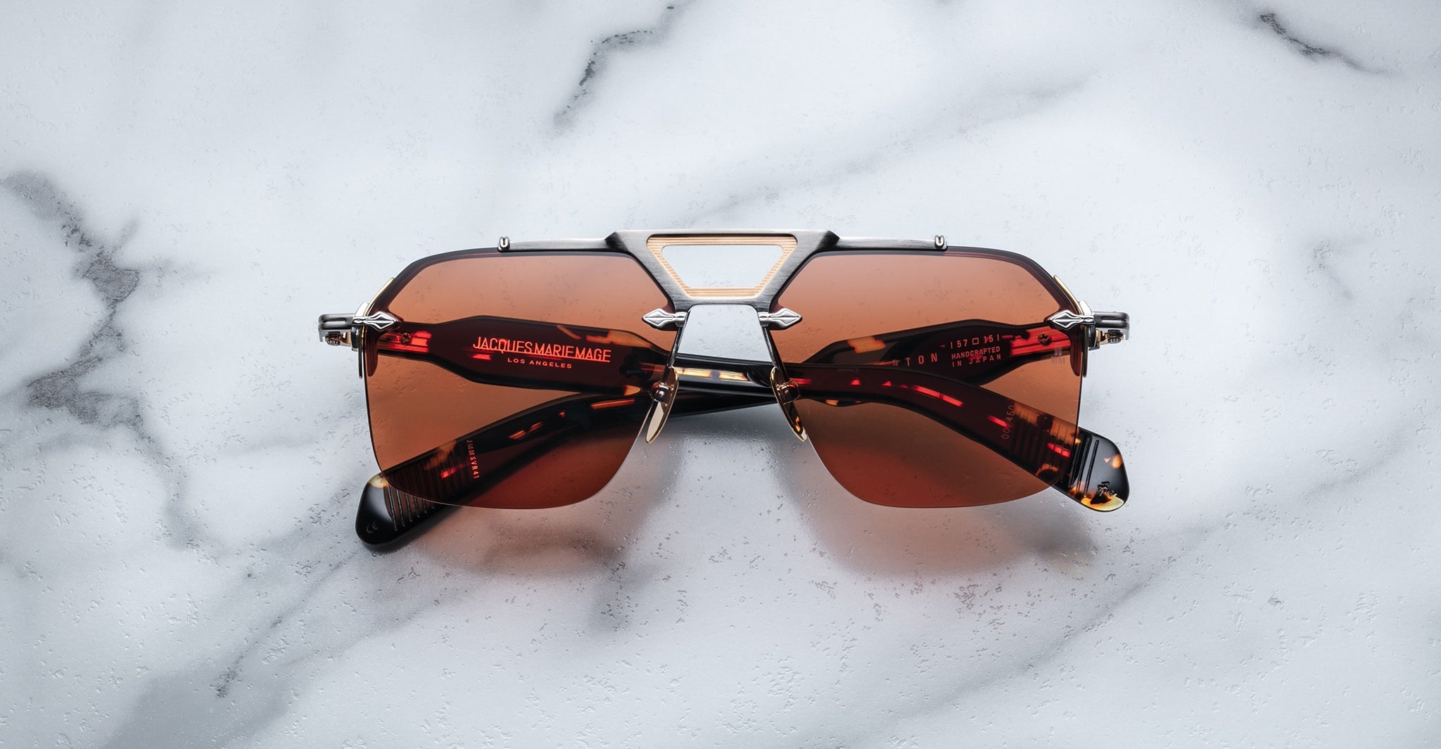 The Jacques Marie Mage Silverton rectangular sunglasses, featuring brown tinted lenses and a silver metal frame, rest on a white marble surface. The brand name and details appear in red text on the arms.