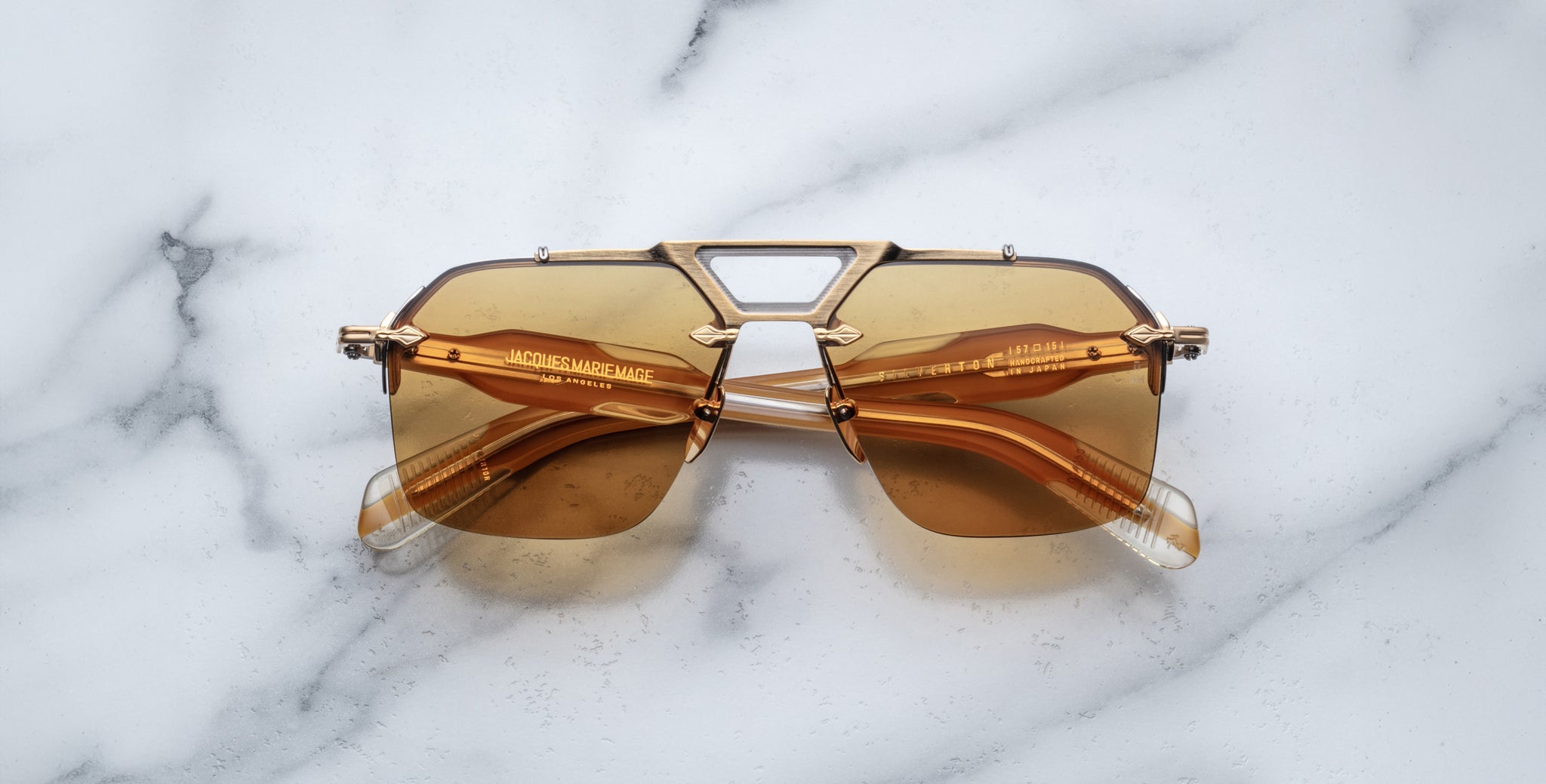 The Jacques Marie Mage Silverton sunglasses, featuring gold rims and brown tinted geometric lenses with visible text on the arms, are displayed on a white marble surface.