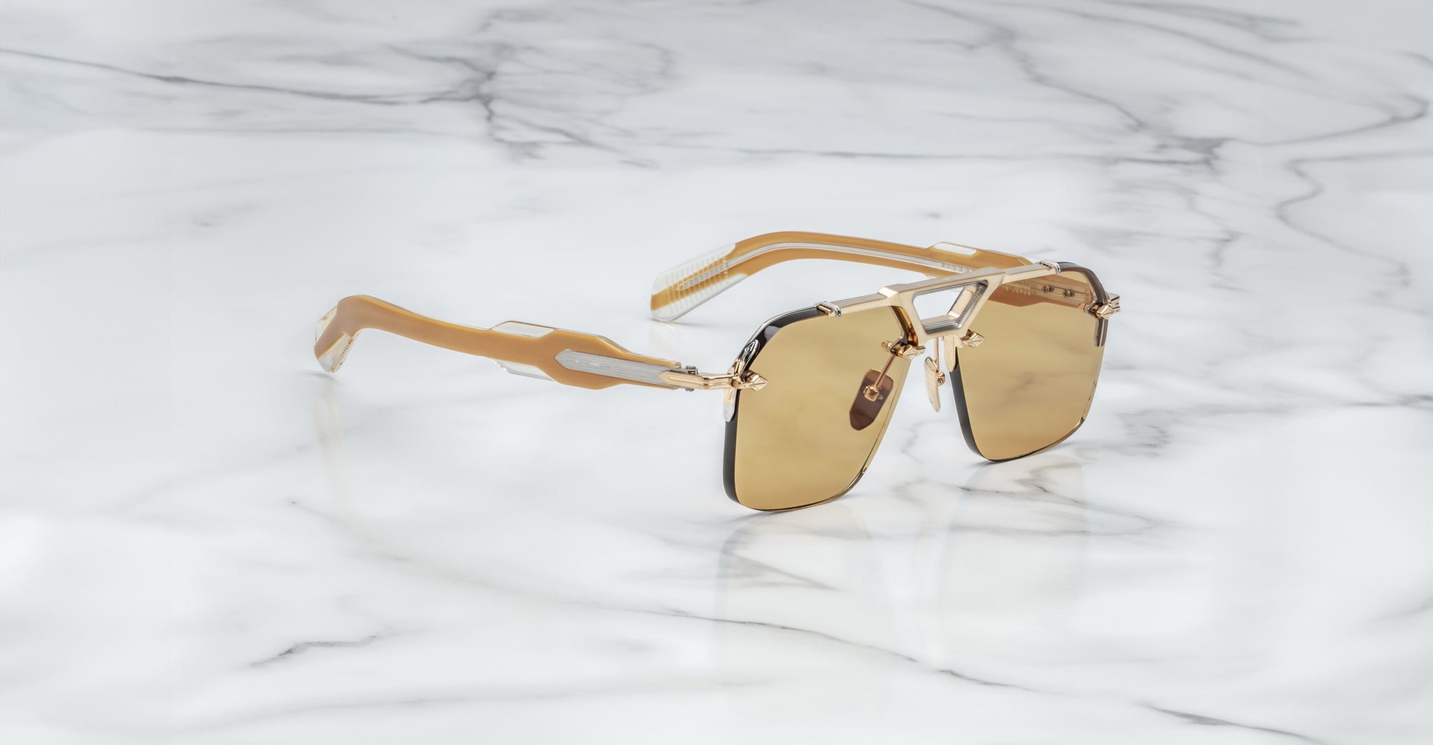 The Jacques Marie Mage Silverton sunglasses, featuring a gold and black frame with metallic geometric accents on the arms and bridge and rectangular brown lenses, rest on a white marble surface.
