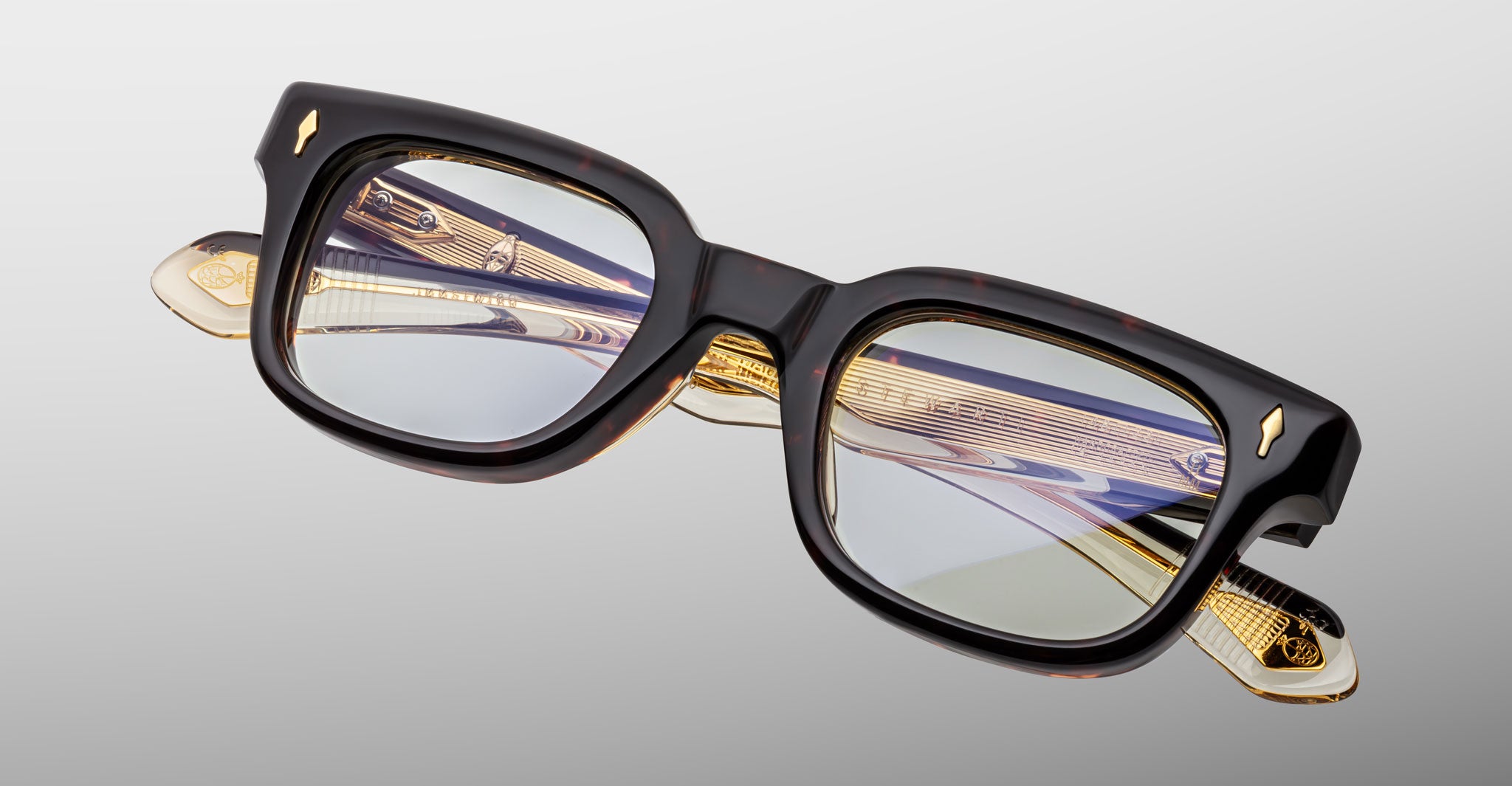 The Jacques Marie Mage Stewart features rectangular, thick-rimmed eyeglasses with dark frames, clear lenses, and yellow-gold metallic temple accents, set against a light gray background.