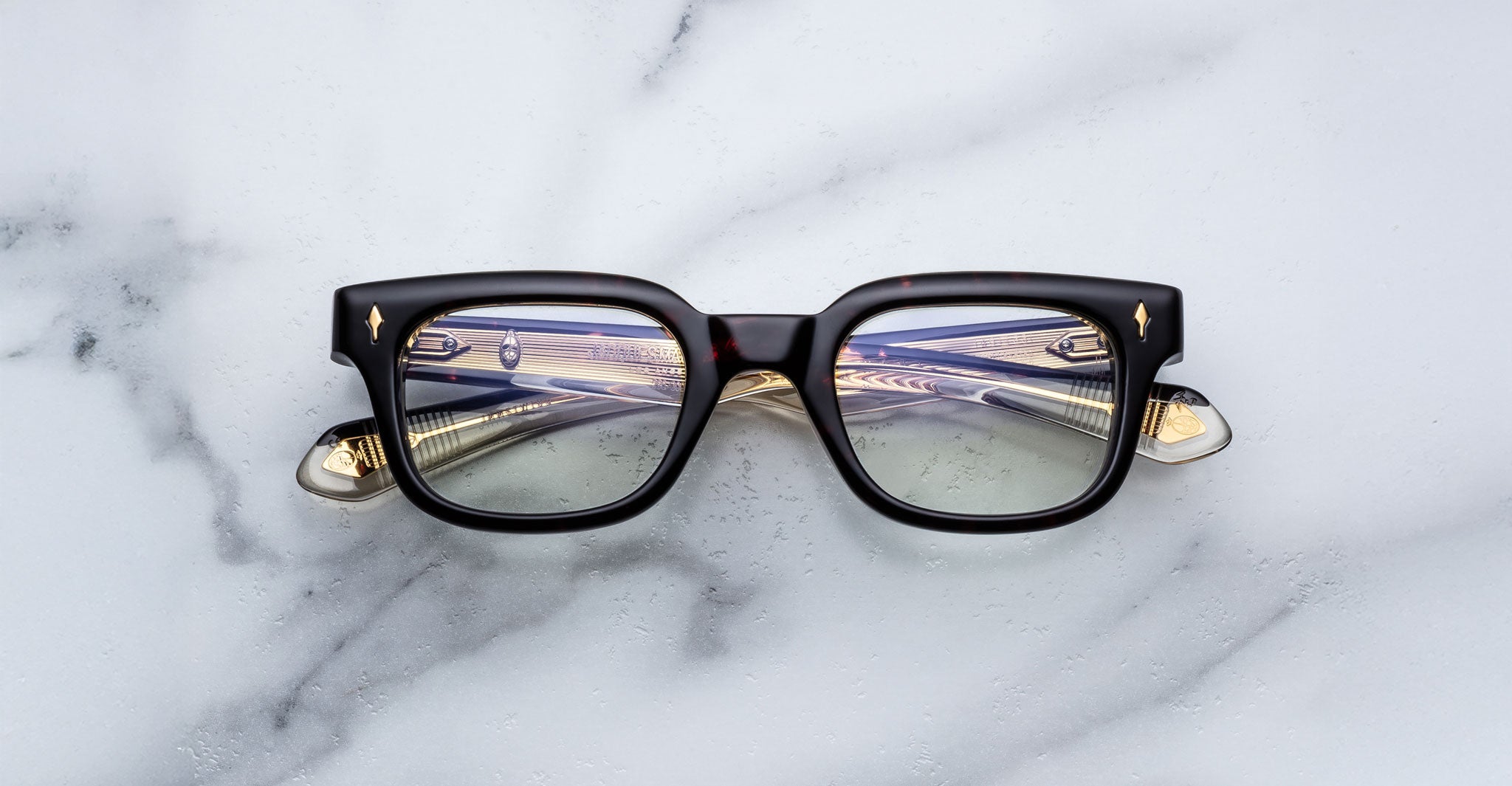 The Jacques Marie Mage Stewart eyeglasses, featuring a black rectangular frame with gold-accented temples, rest on a white marble surface. Striped light reflects off the lenses while parts of the arms show through them.