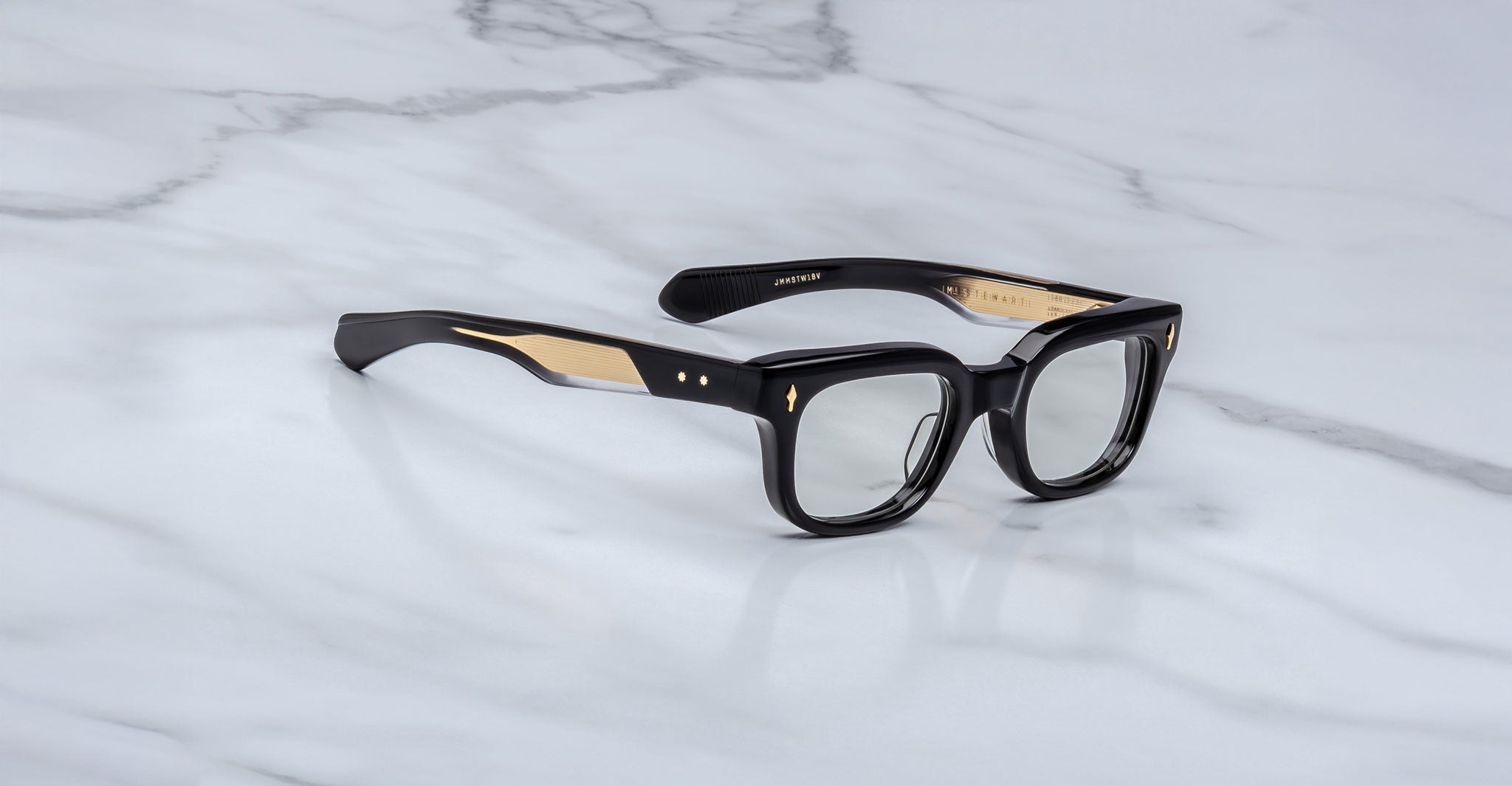 The Jacques Marie Mage Stewart features black and gold rectangular eyeglasses with thick frames and clear lenses, displayed on a white marble surface.