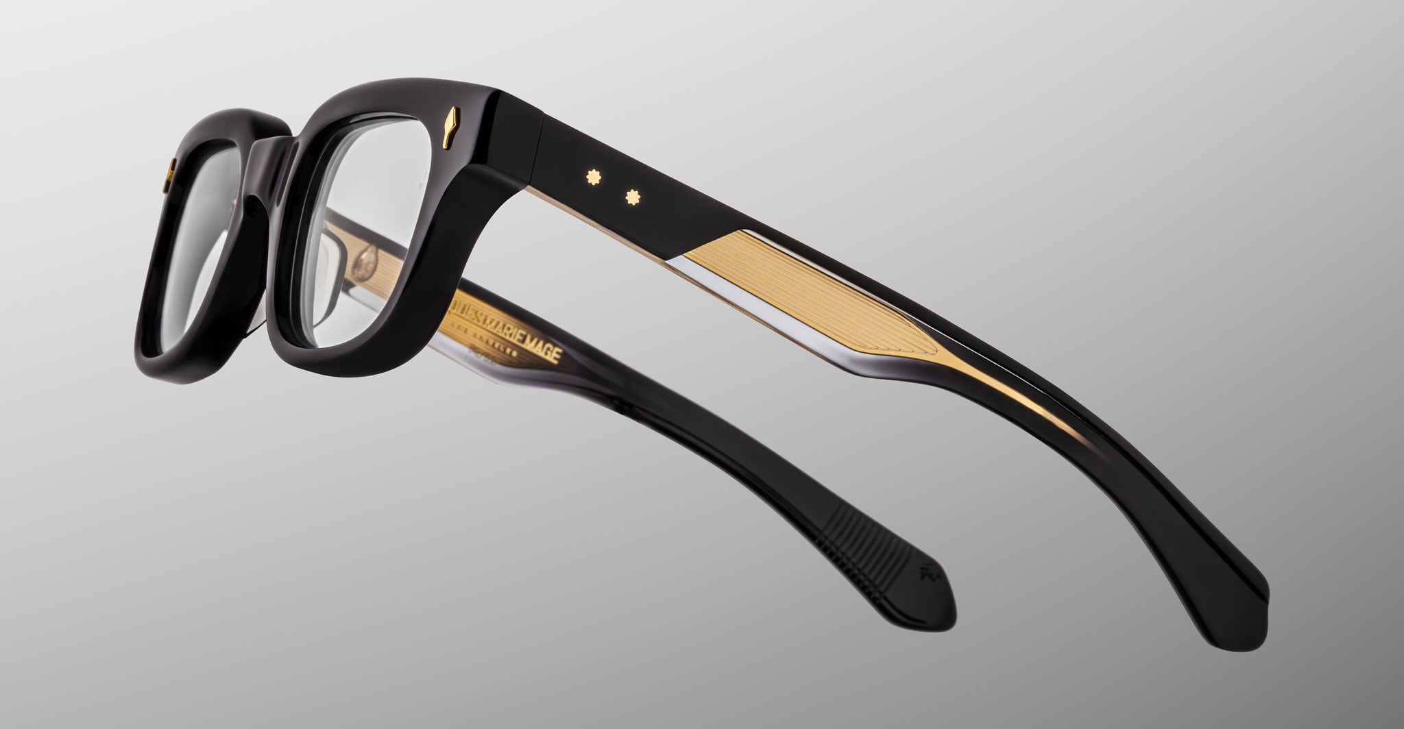The Jacques Marie Mage Stewart eyeglasses feature thick black frames with gold-accented temples, displayed from an angled side view against a light gray background.