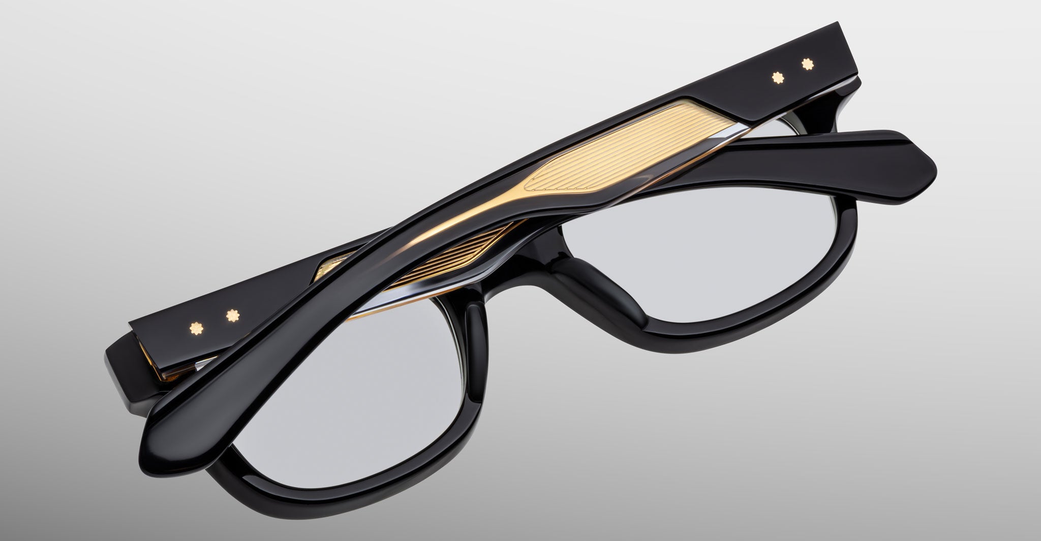 The Jacques Marie Mage Stewart eyeglasses feature a black frame with textured gold accents and small decorative dots on the temples, shown folded on a light gray surface.