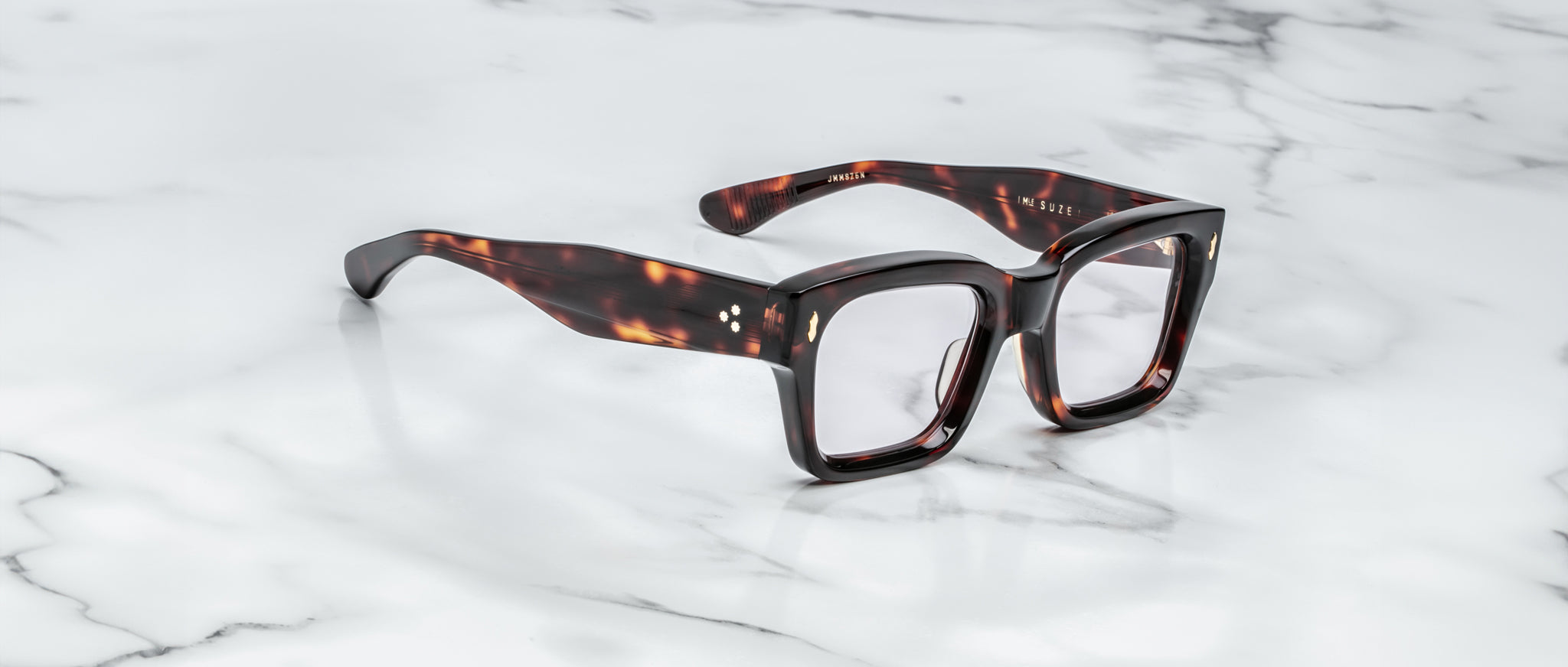 Jacques Marie Mage Suze eyeglasses, featuring a tortoiseshell-patterned rectangular design with thick frames, rest on a smooth white marble surface.