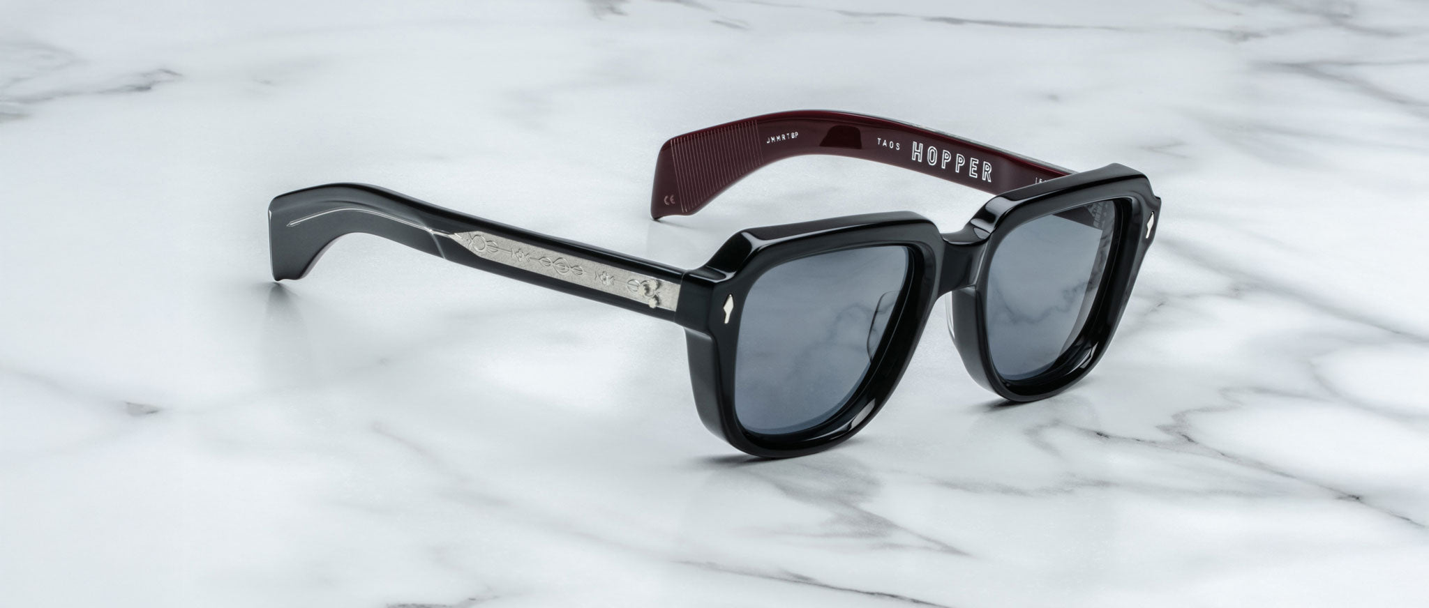 Jacques Marie Mage Taos sunglasses in black and burgundy with dark lenses, silver accents on the arms, and "HOPPER" inside the left arm, shown on a white marble surface.