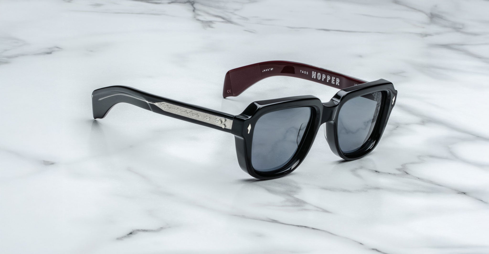 Jacques Marie Mage Taos sunglasses in black and burgundy with dark lenses, silver accents on the arms, and "HOPPER" inside the left arm, shown on a white marble surface.