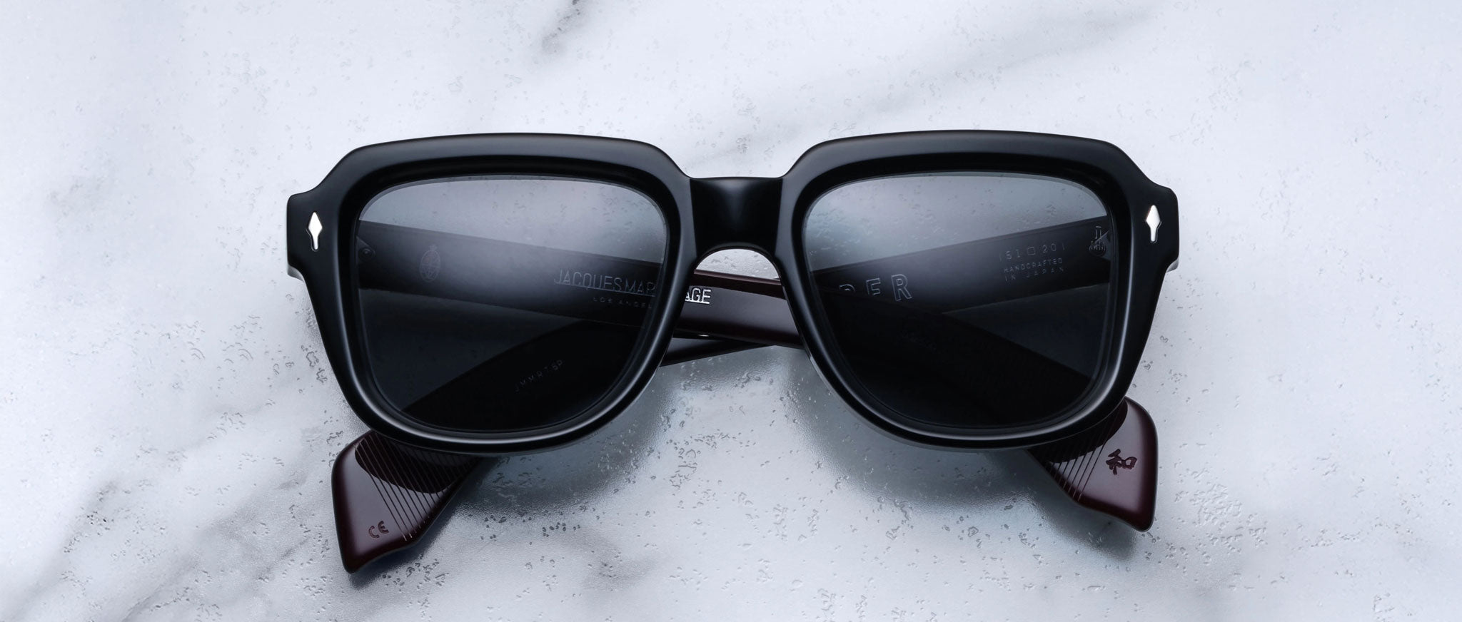 A pair of Jacques Marie Mage Taos black rectangular sunglasses with dark lenses and thick frames rests on a white marble surface.