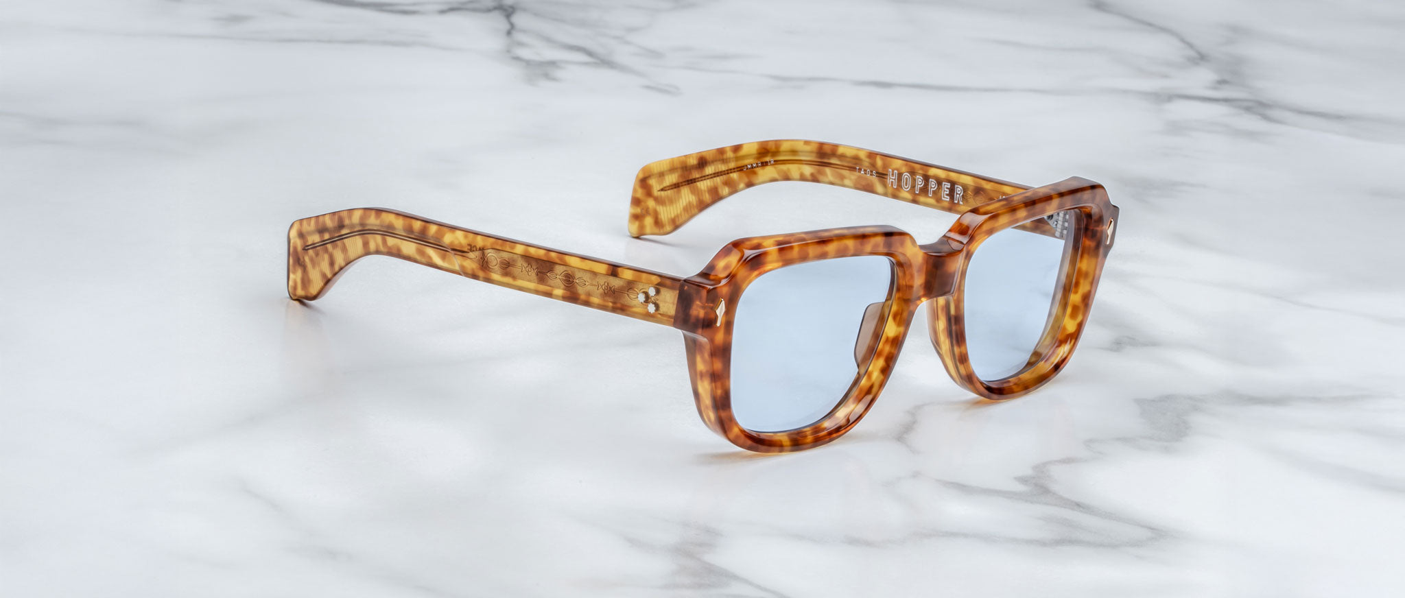 A pair of Jacques Marie Mage Taos square tortoiseshell sunglasses with blue-tinted lenses rests on a white marble surface.
