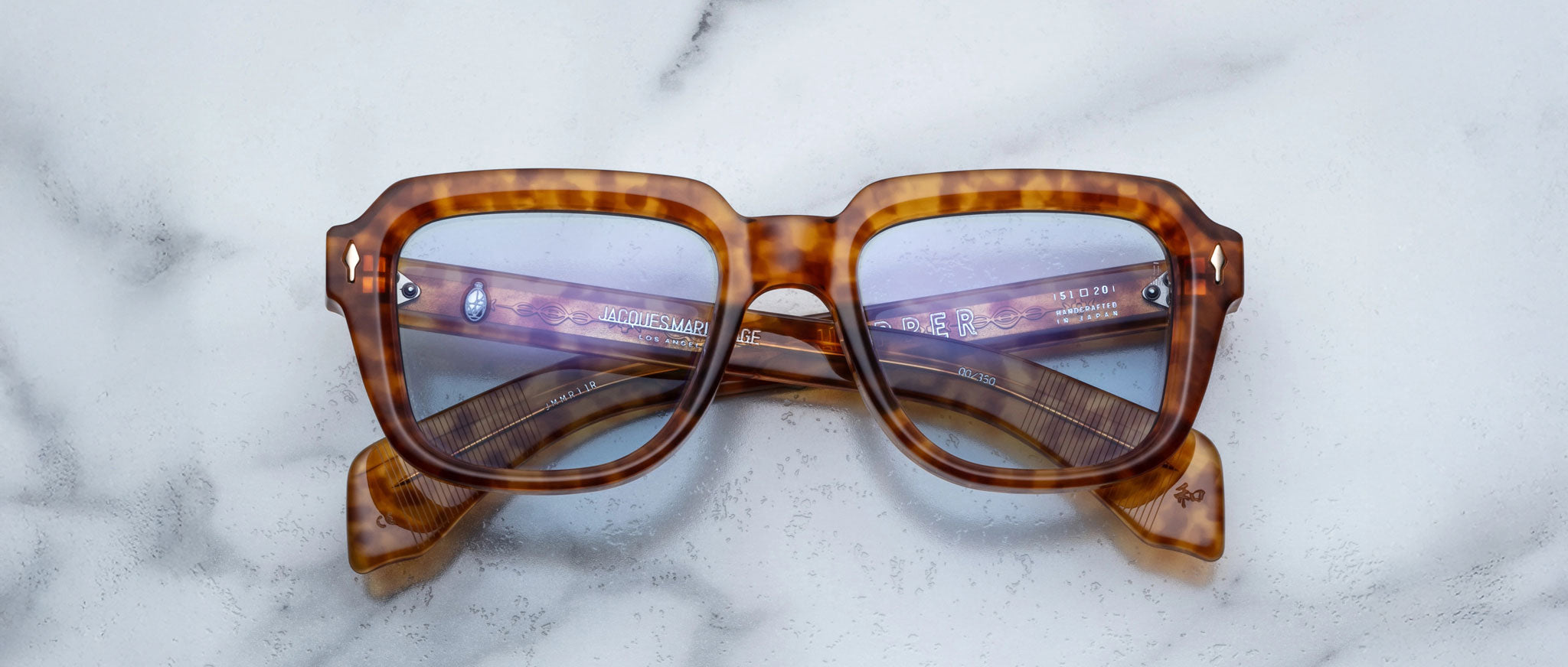 Jacques Marie Mage Taos tortoiseshell square eyeglasses with blue-tinted lenses are displayed with crossed arms on a light marble surface.