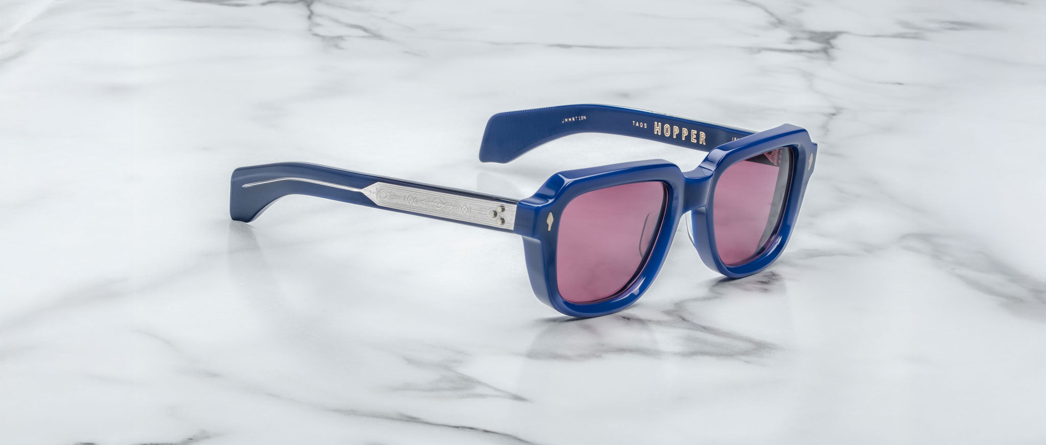 Jacques Marie Mage Taos sunglasses with blue frames and pink-tinted lenses rest on a white marble surface. "HOPPER" is printed in gold on the inner left arm.