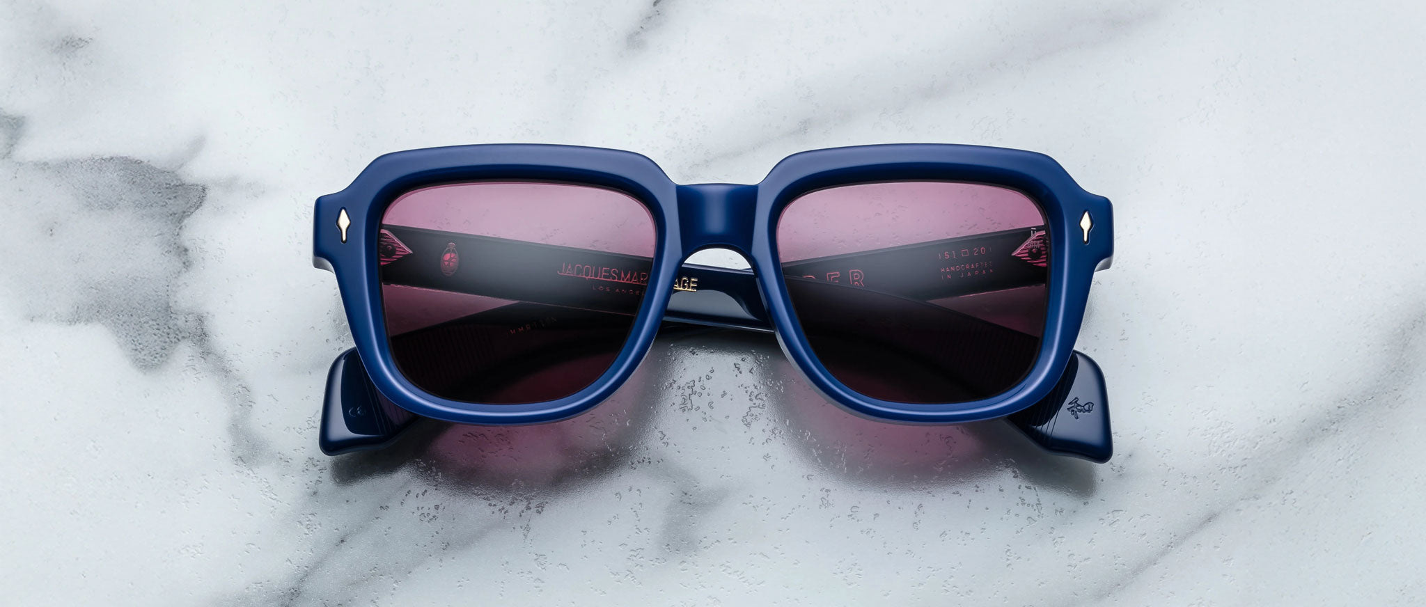 The Jacques Marie Mage Taos sunglasses, featuring blue frames and pink-tinted lenses, are displayed on a white marble surface in an overhead view.