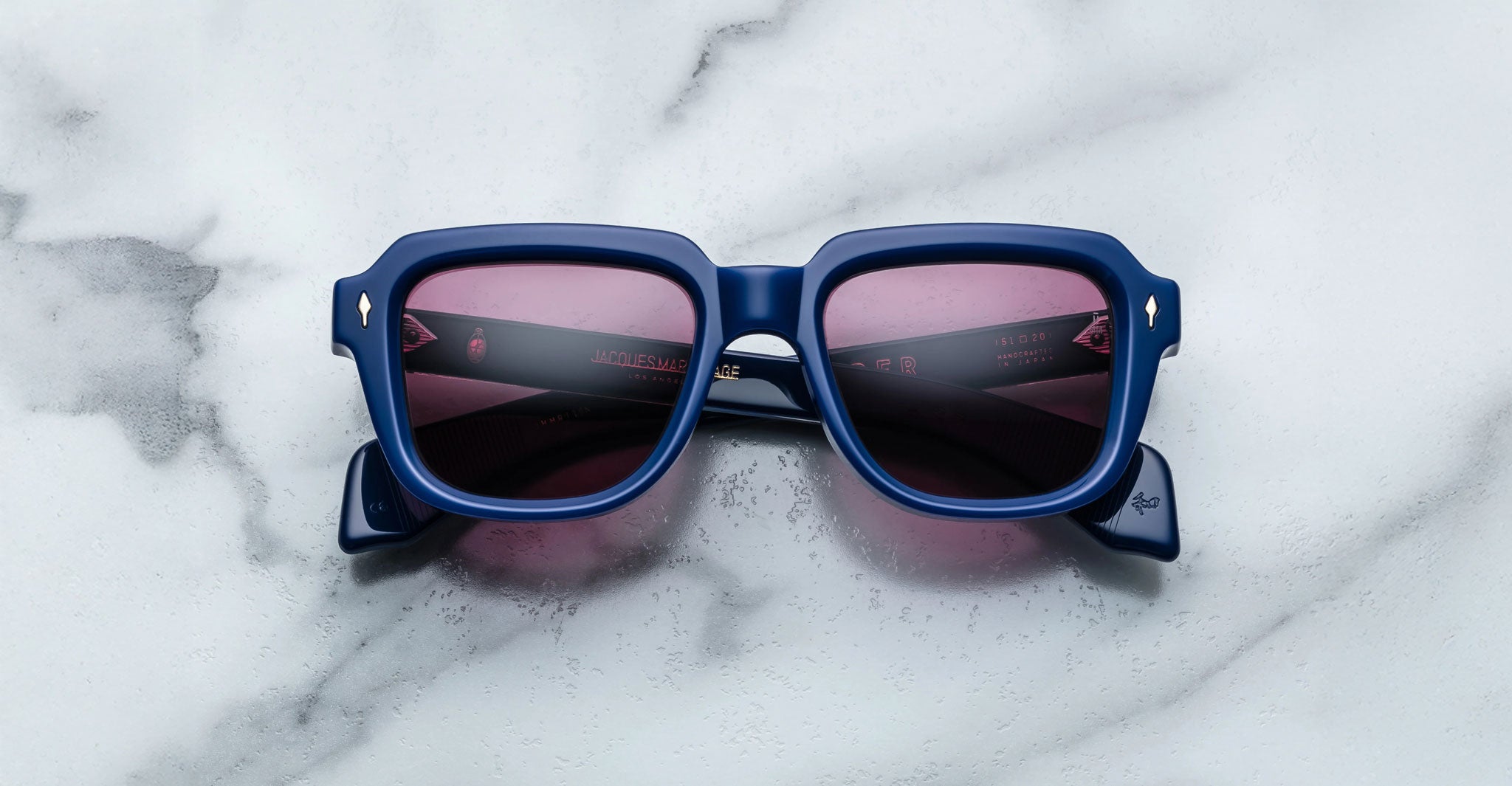 The Jacques Marie Mage Taos sunglasses, featuring blue frames and pink-tinted lenses, are displayed on a white marble surface in an overhead view.