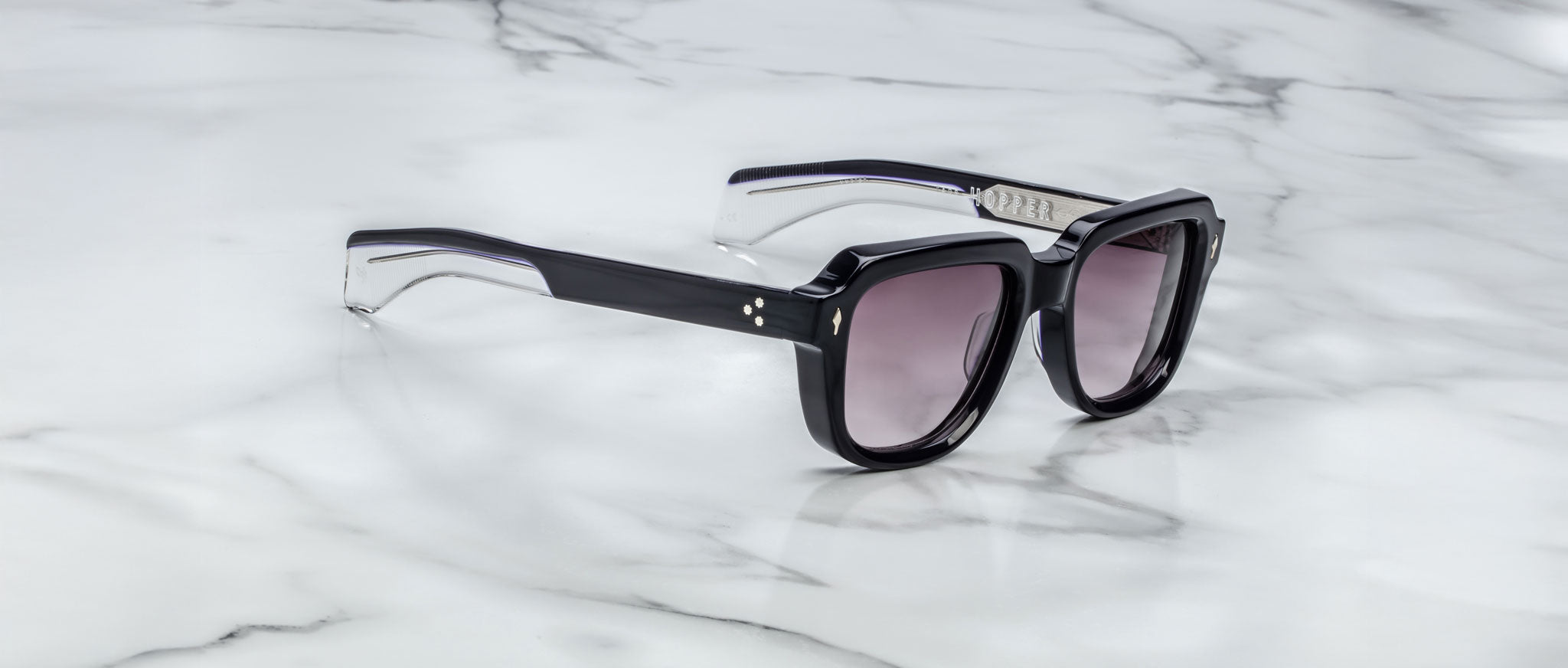 The Jacques Marie Mage Taos sunglasses feature thick black frames, gradient tinted lenses, and silver accents near the hinges. They are pictured resting on a white marble surface.