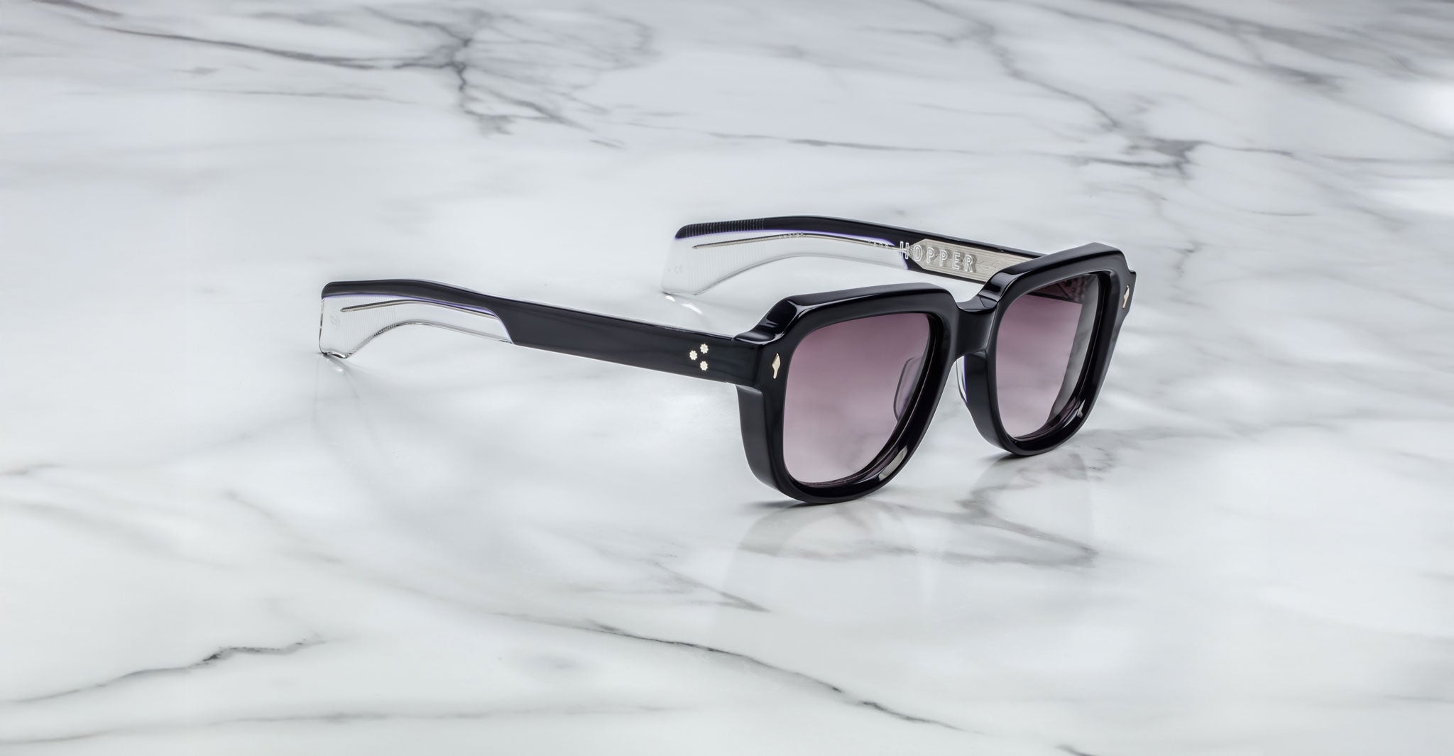 The Jacques Marie Mage Taos sunglasses feature thick black frames, gradient tinted lenses, and silver accents near the hinges. They are pictured resting on a white marble surface.