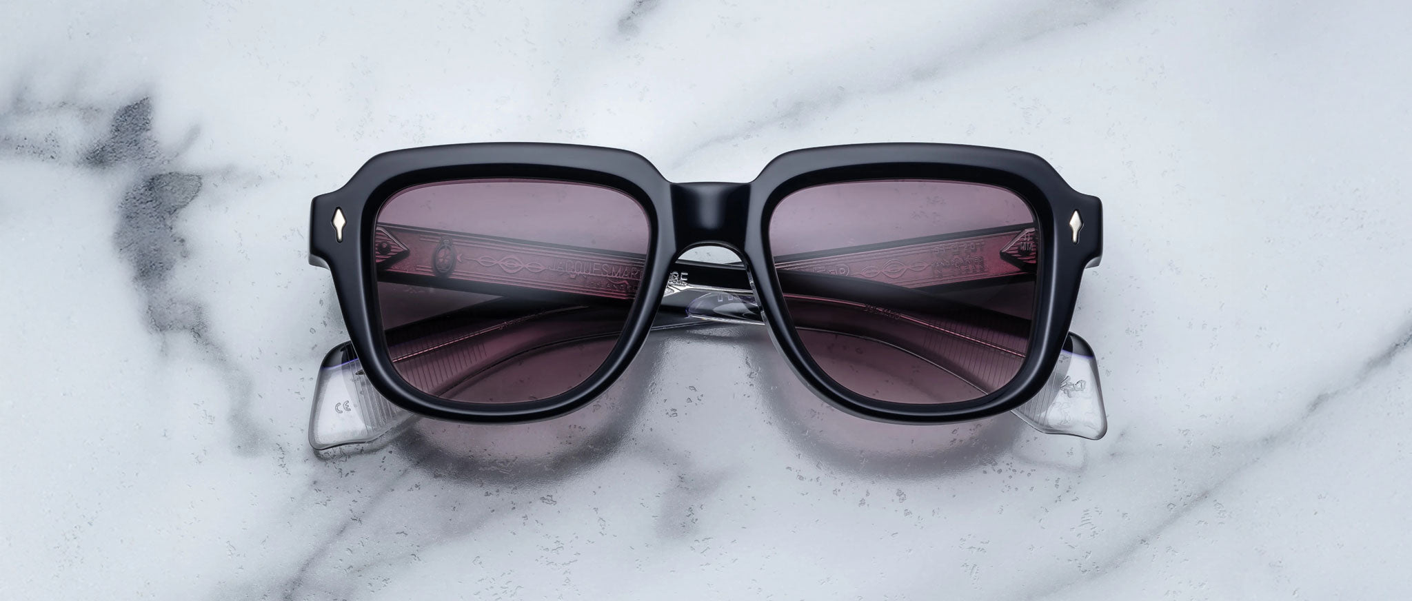 The Jacques Marie Mage Taos sunglasses, featuring black square frames and dark tinted lenses, are displayed on a white marble surface with gray veining.