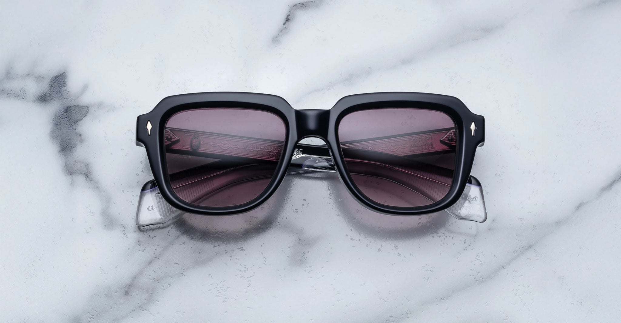The Jacques Marie Mage Taos sunglasses, featuring black square frames and dark tinted lenses, are displayed on a white marble surface with gray veining.