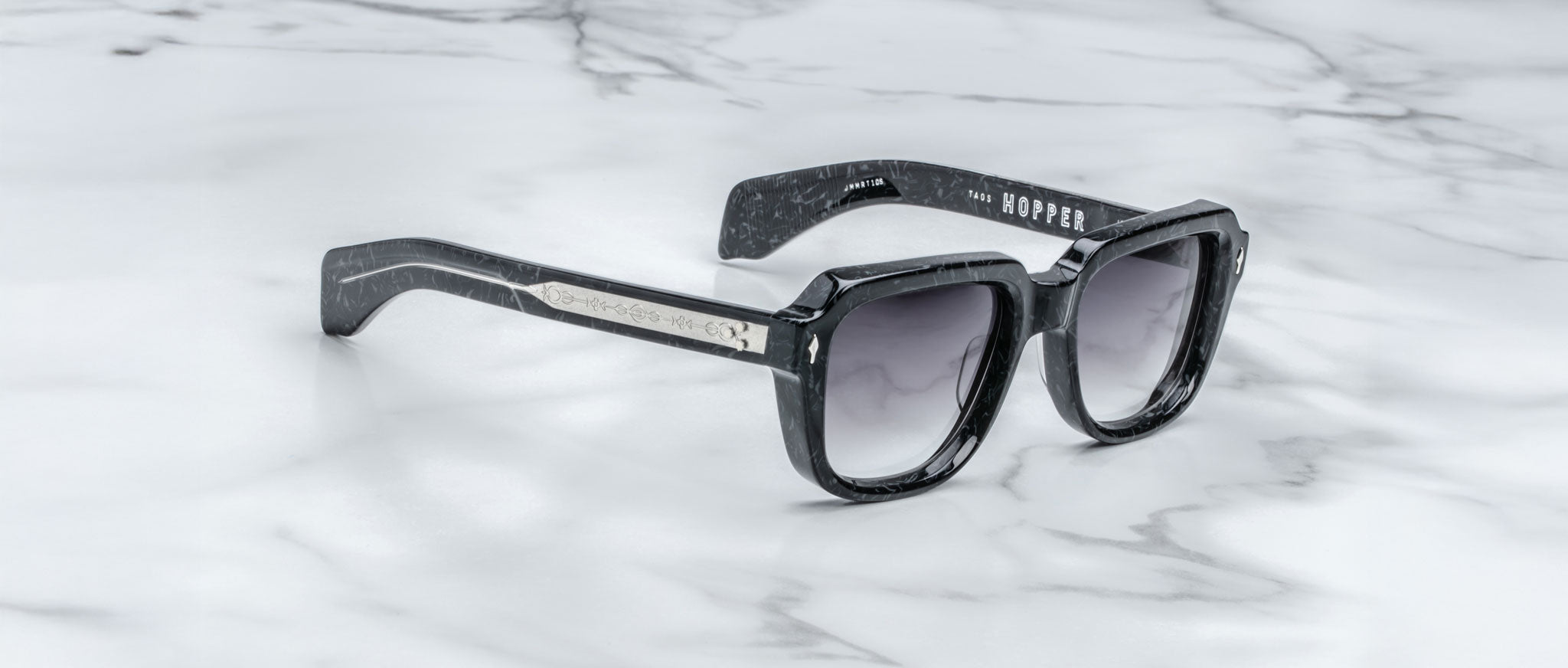 Jacques Marie Mage Taos black square-framed sunglasses with dark gradient lenses sit on a white marble surface. Subtle silver details accent the frame, and “JACQUES MARIE MAGE - TAOS” is printed inside the left temple arm.