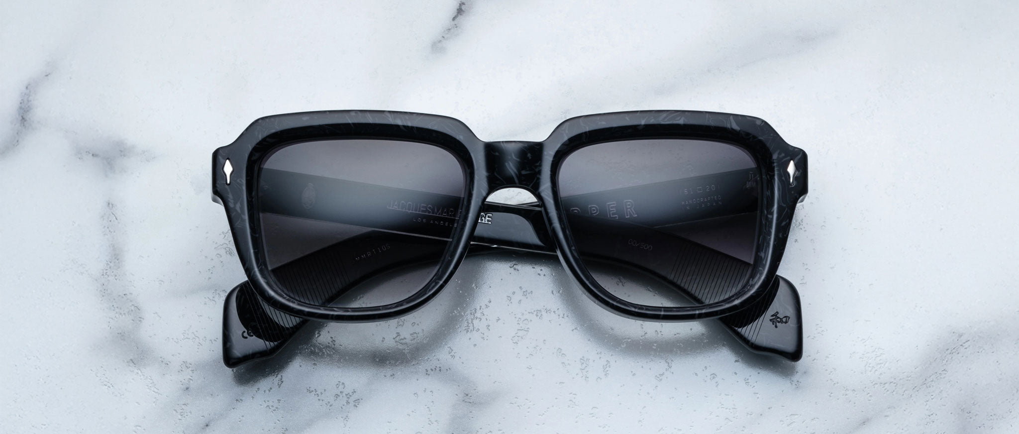 A pair of Jacques Marie Mage Taos black square sunglasses with dark lenses rests on a white marble surface. The crossed arms and silver accents on the upper corners highlight the elegant design.
