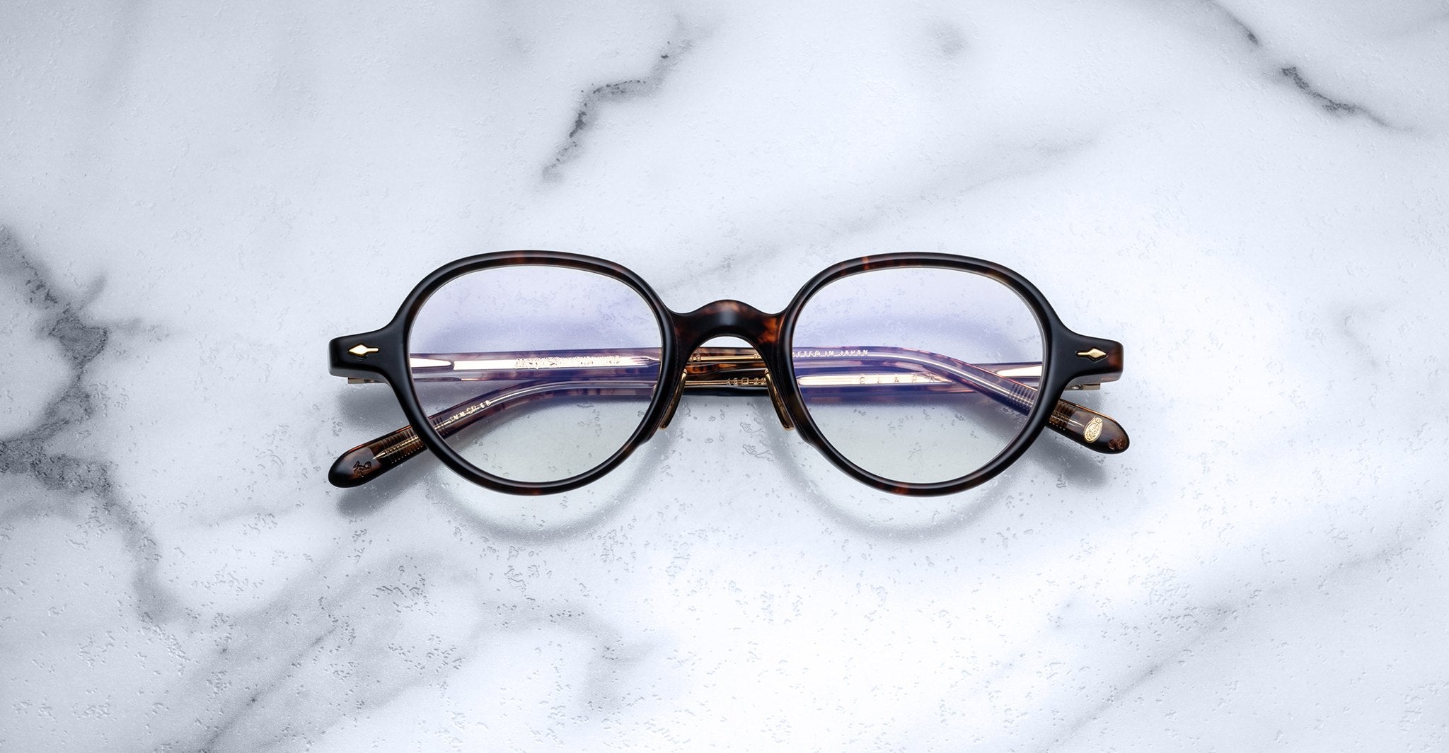 Jacques Marie Mage Clark round tortoiseshell eyeglasses with folded temples rest on white marble—a midcentury-inspired optical frame by Jacques Marie Mage, blending classic design with timeless style.