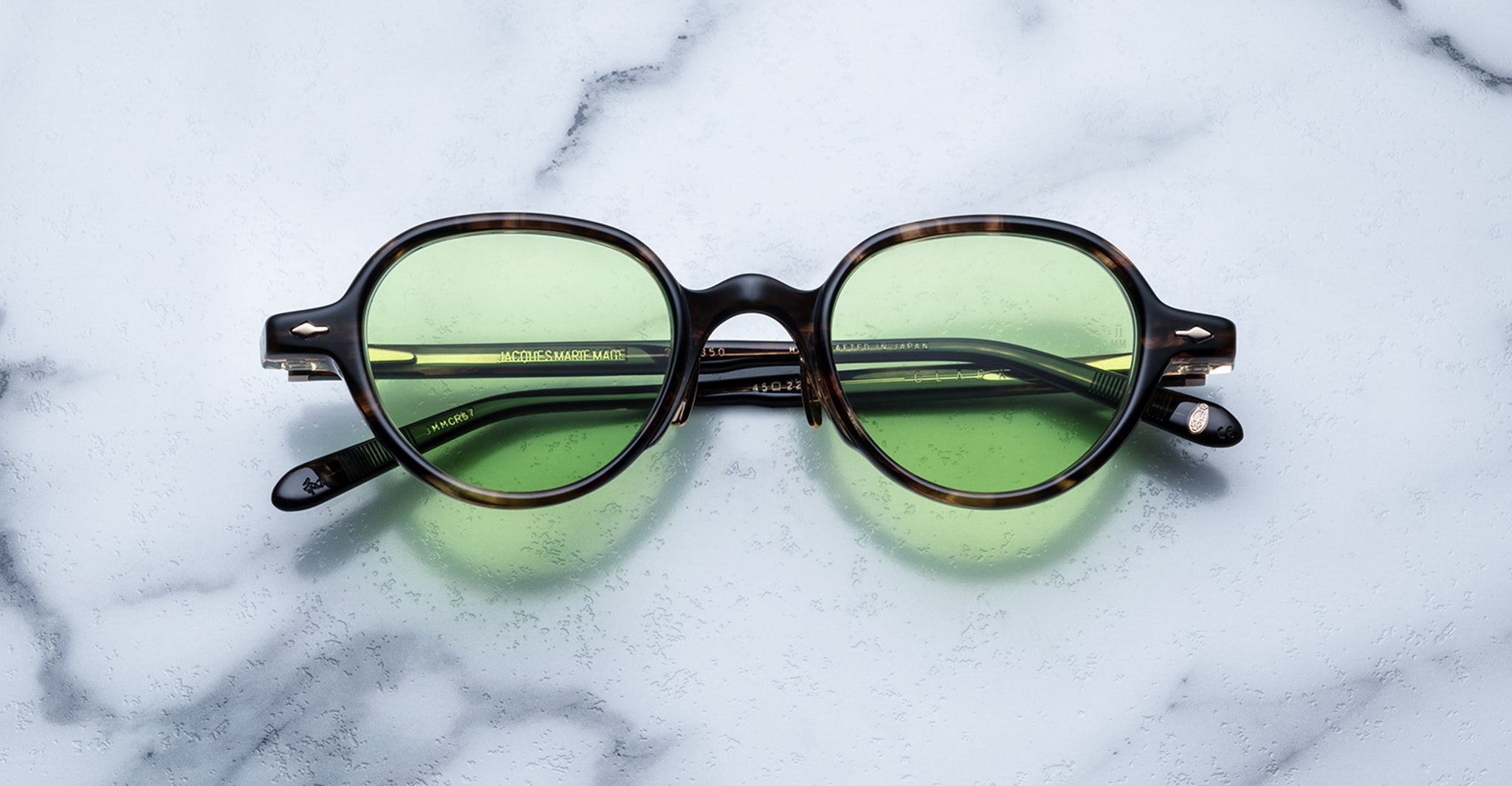 Jacques Marie Mage Clark eyeglasses with green-tinted lenses and dark rims rest on white marble, highlighting the brand's signature midcentury-inspired elegance.