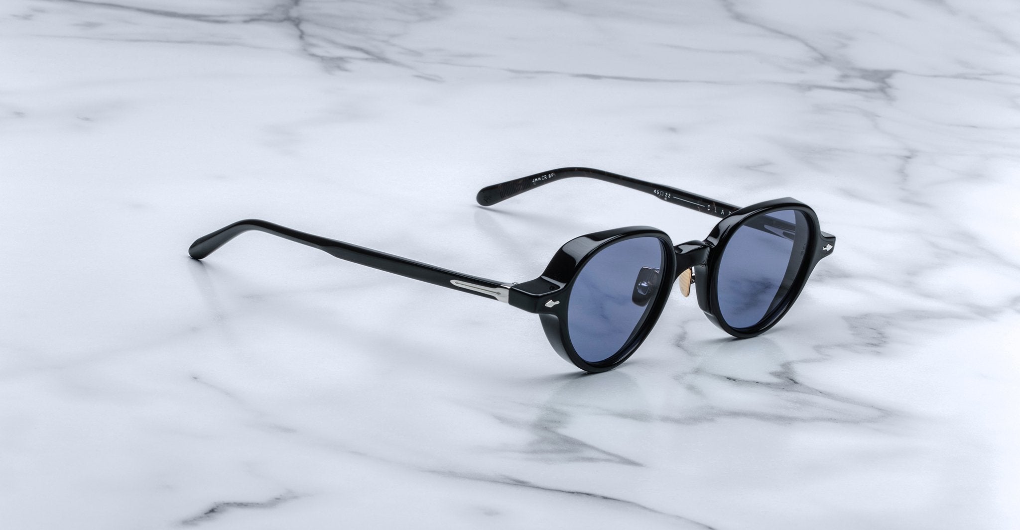 The Jacques Marie Mage Clark sunglasses by Jacques Marie Mage feature black round frames with dark lenses, inspired by midcentury style, displayed on a white marble surface.