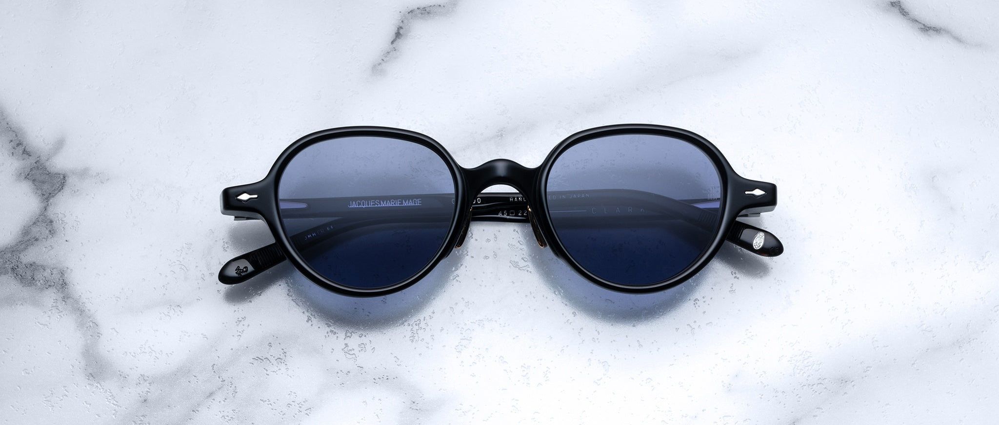 The Jacques Marie Mage Clark sunglasses feature round, black frames and dark lenses with midcentury-inspired design, shown on a white marble surface.