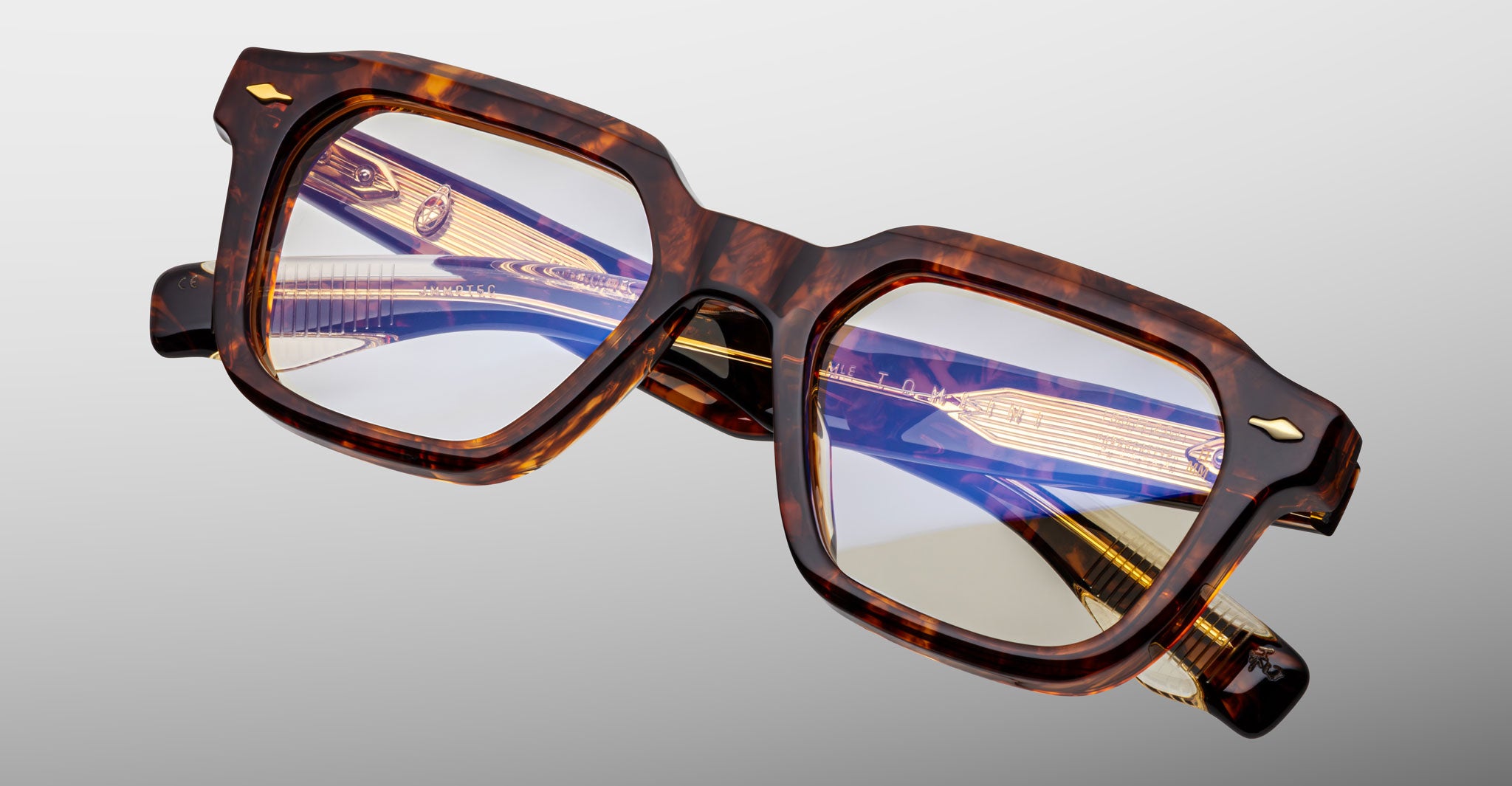 The Jacques Marie Mage Tomlin eyeglasses feature thick, square tortoiseshell frames with slightly reflective lenses and gold and purple detailing on the arms, displayed on a light gray background.