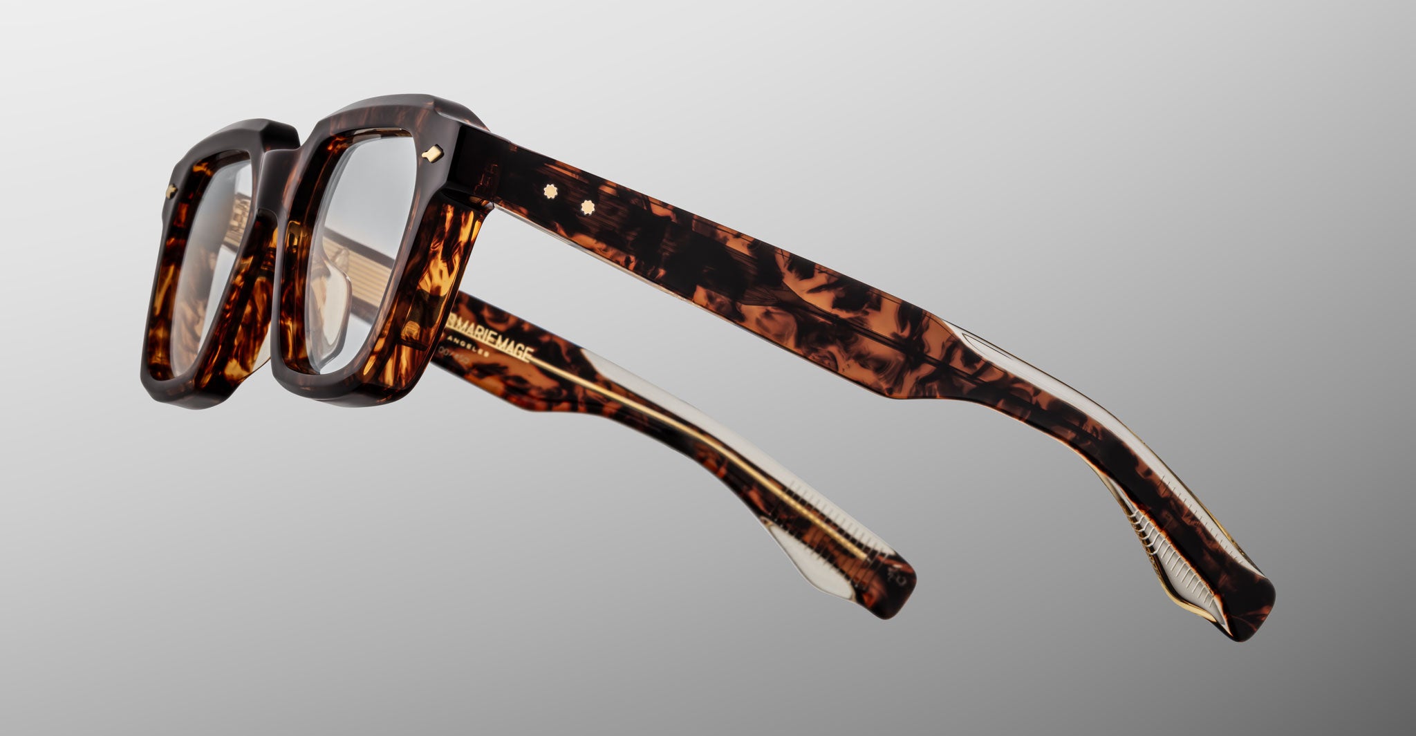 The Jacques Marie Mage Tomlin features thick, rectangular tortoiseshell frames with visible arms and hinges, displayed against a neutral gray background.