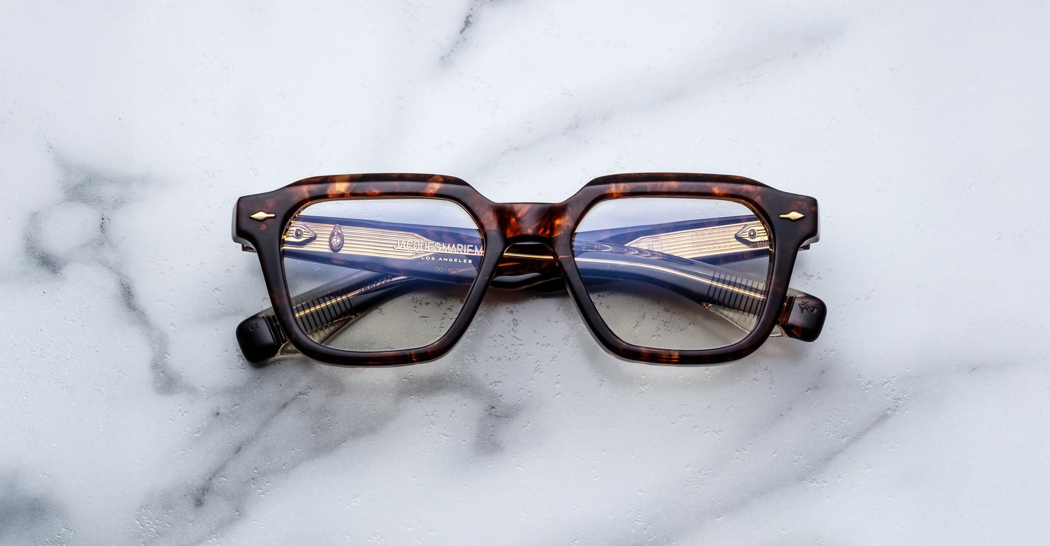 A pair of Jacques Marie Mage Tomlin tortoiseshell rectangular eyeglasses rests on a white marble surface, with the lenses reflecting a metallic object featuring horizontal lines.