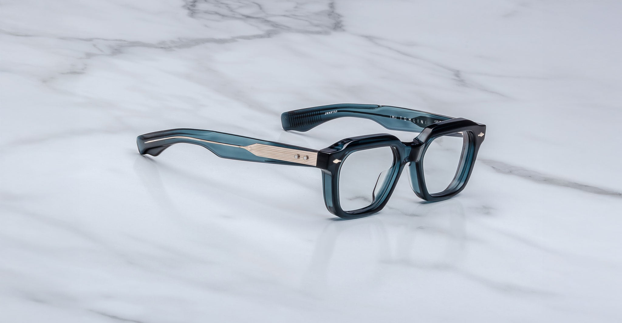 The Jacques Marie Mage Tomlin eyeglasses feature thick, dark teal frames and wide arms with metallic gold accents, displayed on a white marble surface.