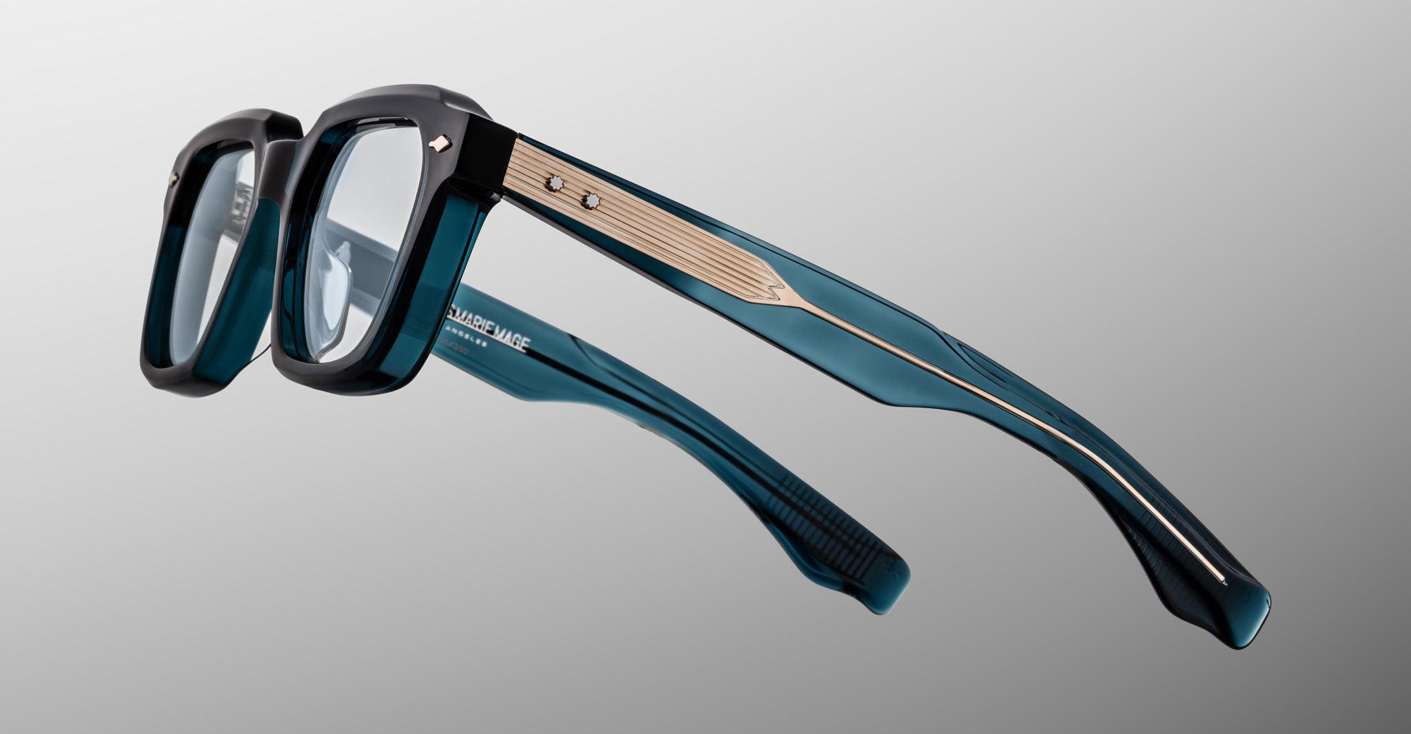 Jacques Marie Mage Tomlin eyeglasses feature teal and black thick rectangular frames, clear lenses, gold metal temple accents, and visible hinge screws, displayed on a plain gradient background.