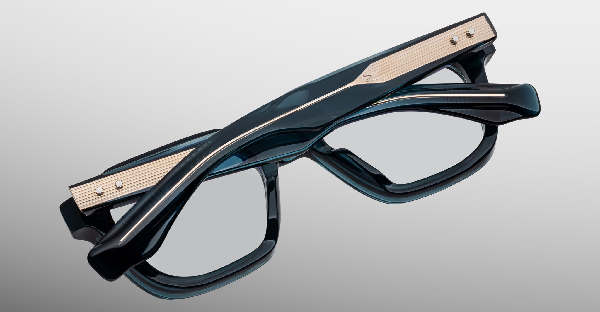 A close-up of Jacques Marie Mage Tomlin black and gold rectangular eyeglasses with thick frames, shown folded on a light grey gradient background.