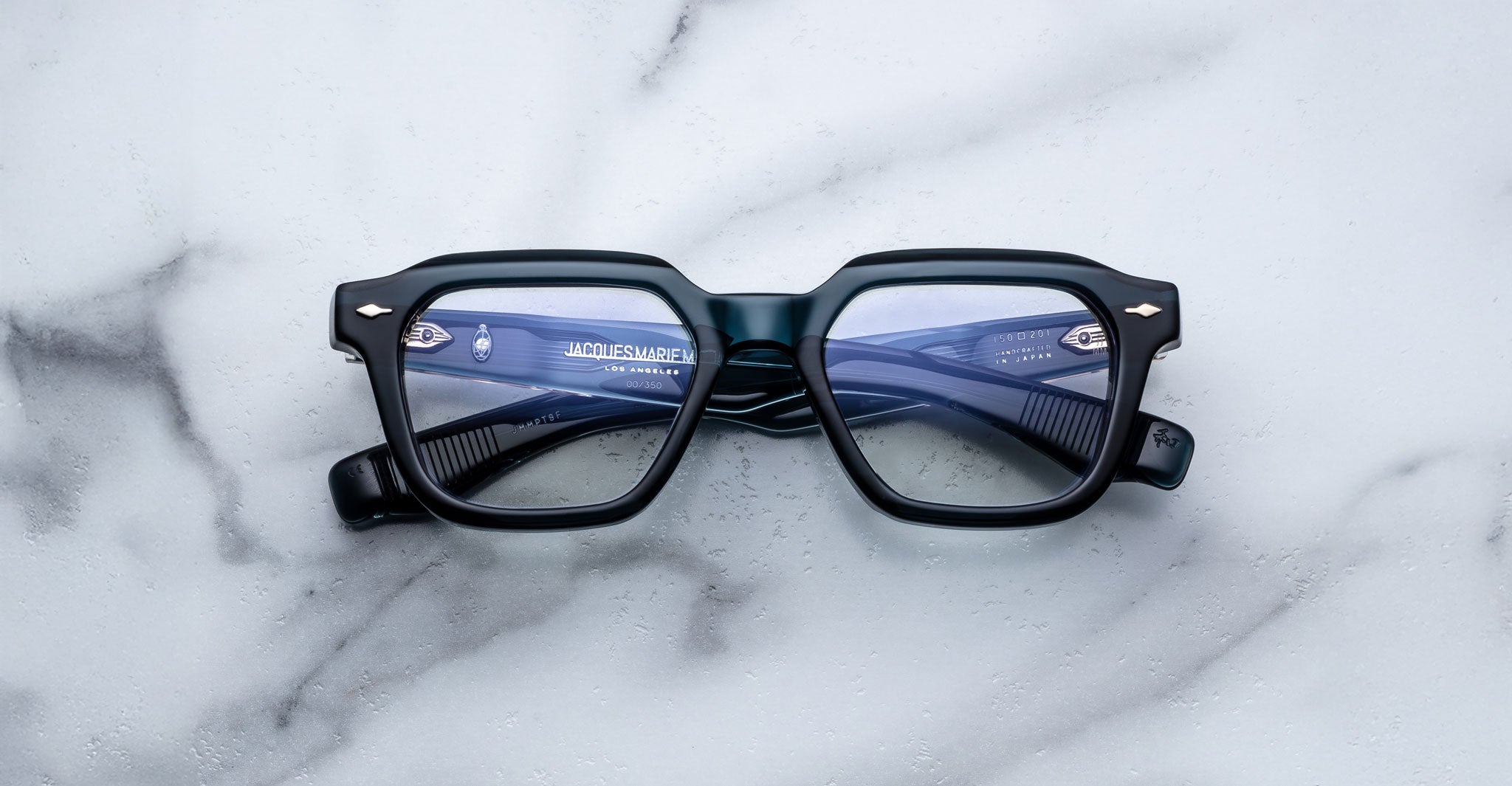 A pair of Jacques Marie Mage Tomlin rectangular black eyeglasses with blue-tinted lenses rests on a white marble surface, showcasing the temples and branding through the lenses.