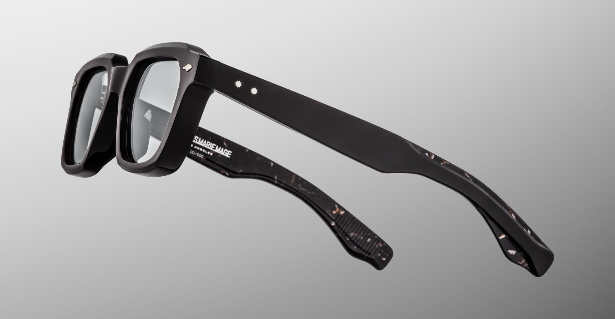 A side view of the Jacques Marie Mage Tomlin eyeglasses showcases their black rectangular thick frames, subtle marbled pattern on the inner temples, and small decorative accents near the hinges, set against a gray background.